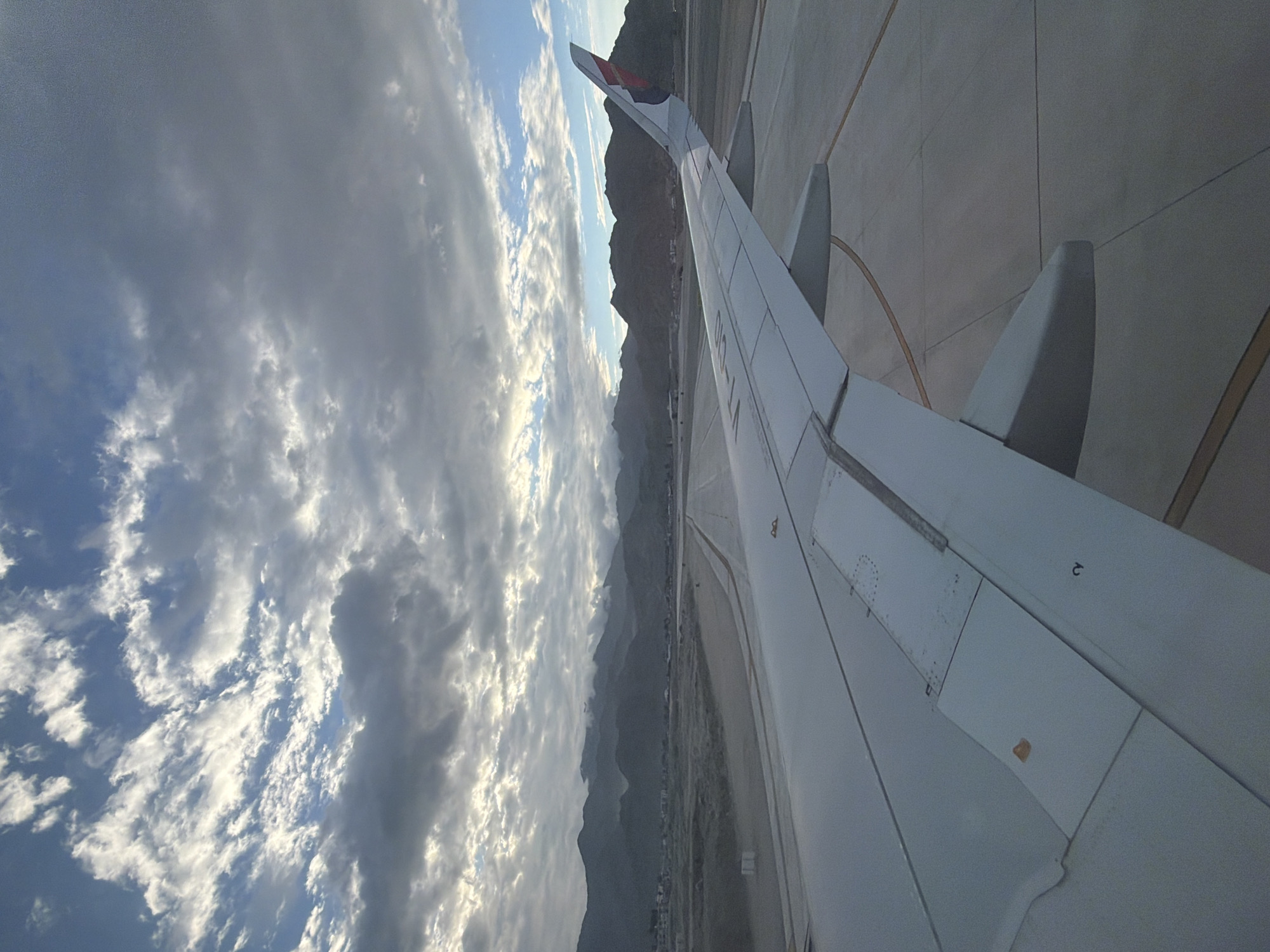 The image captures a view from an airplane wing, likely on the tarmac at Kushok Bakula Rimpochee Airport in Spituk, India. The foreground is dominated by the broad, white wing of the aircraft, which extends diagonally across the frame. The wing features visible panel lines and a small, orange-brown marking. To the right of the wing, a section of the airport tarmac is visible, marked with yellow lines. In the mid-ground, a runway stretches into the distance, flanked by barren, sandy terrain. Beyond the runway, a hazy view of mountains rises against a backdrop of a cloudy sky. The clouds are a mix of dark grays and bright whites, with patches of blue sky visible. The overall lighting suggests it might be early morning or late afternoon, given the diffused sunlight. There are no people or moving vehicles visible in the image.