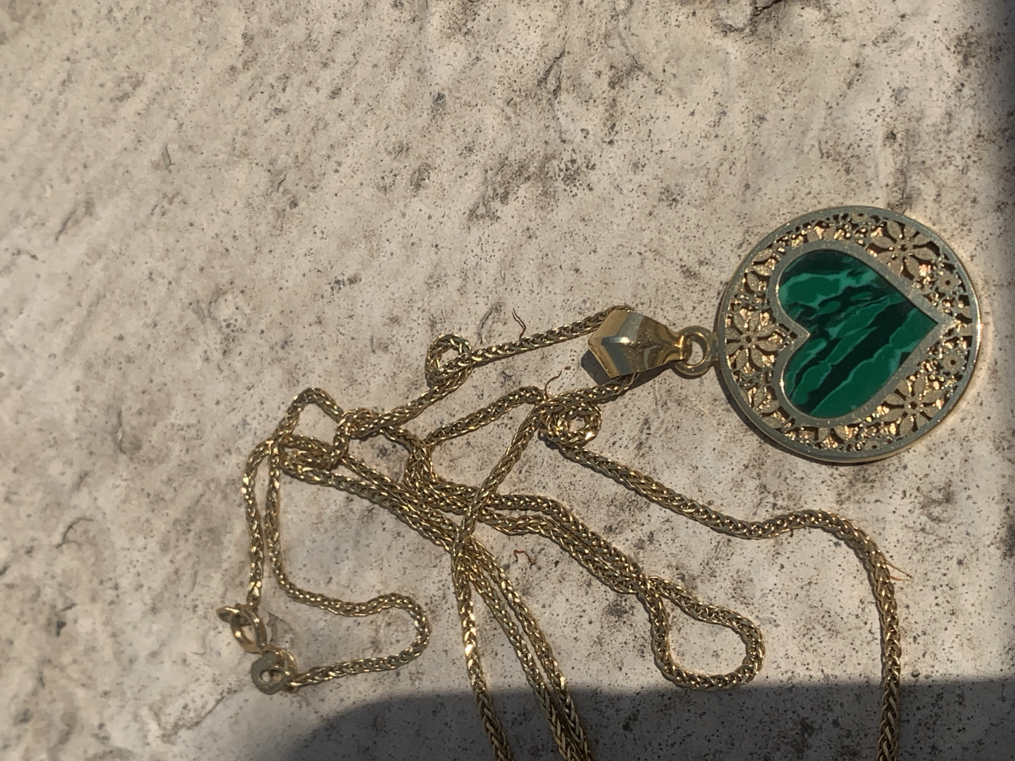 A close-up shot shows a gold necklace with a heart-shaped malachite pendant lying on a textured, light-colored surface. The pendant is circular and features an intricate gold filigree border surrounding the green malachite stone, which displays characteristic banding patterns. The gold chain is draped loosely across the surface, with a lobster clasp visible near the top left of the image. The lighting suggests it is outdoors, casting a shadow from the chain onto the surface. There are no people or actions depicted. The surface appears to be a stone or concrete material, possibly an outdoor patio or pathway. The location of Khazar Raion, Azerbaijan, is not visually represented in the image itself.