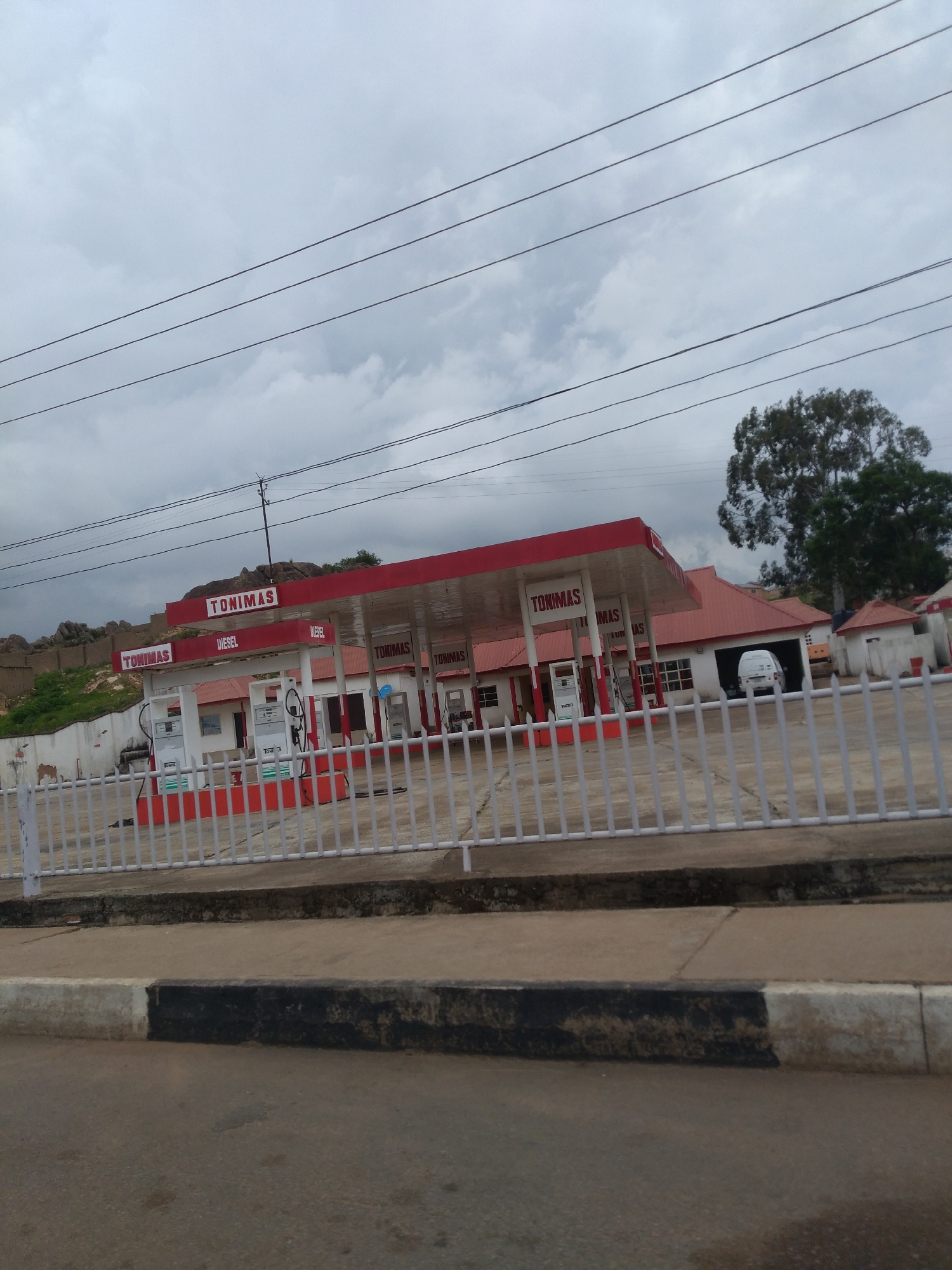 A Tonimas gas station is visible on a cloudy day. The gas station has a red roof and white walls, with "TONIMAS" signage prominently displayed on the canopy and the building. There are several fuel pumps under the canopy, and a white van is parked inside a garage. A white fence runs along the front of the gas station, separating it from the road. In the background, there are rocky hills and a large tree. The scene appears to be in Jos, Nigeria, given the presence of the Tonimas gas station brand, which is common in Nigeria.
