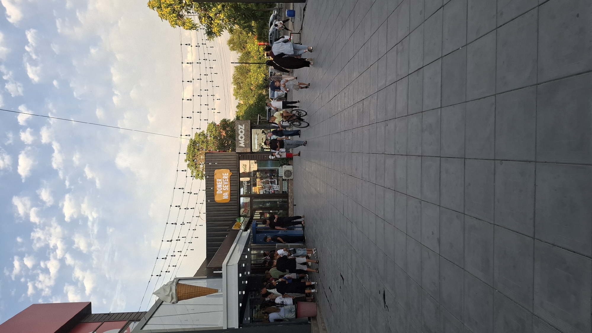 The image captures a street scene in Shymkent, Kazakhstan, likely during daylight hours given the bright sky and visible sunlight. The scene features a pedestrian walkway paved with square tiles, running alongside a row of modern, container-like businesses. A string of decorative lights hangs overhead, suggesting an evening or festive atmosphere, though the bright sky contradicts this.
Several people are visible throughout the scene. Some are walking along the paved area, while others are gathered in front of the businesses, possibly queuing or socializing. One individual is seen riding a bicycle. The businesses appear to be food establishments; one prominently displays the text "DONER NA SEVERE" on an orange sign and another has a "MOOZ" logo. Other visible text includes "Coca-Cola". A large, decorative ice cream cone is perched on top of one of the structures, reinforcing the food-related nature of the businesses. The sky is a bright blue with scattered white clouds, and lush green trees are visible in the background, suggesting a pleasant outdoor environment. The overall impression is one of a lively, urban commercial area.