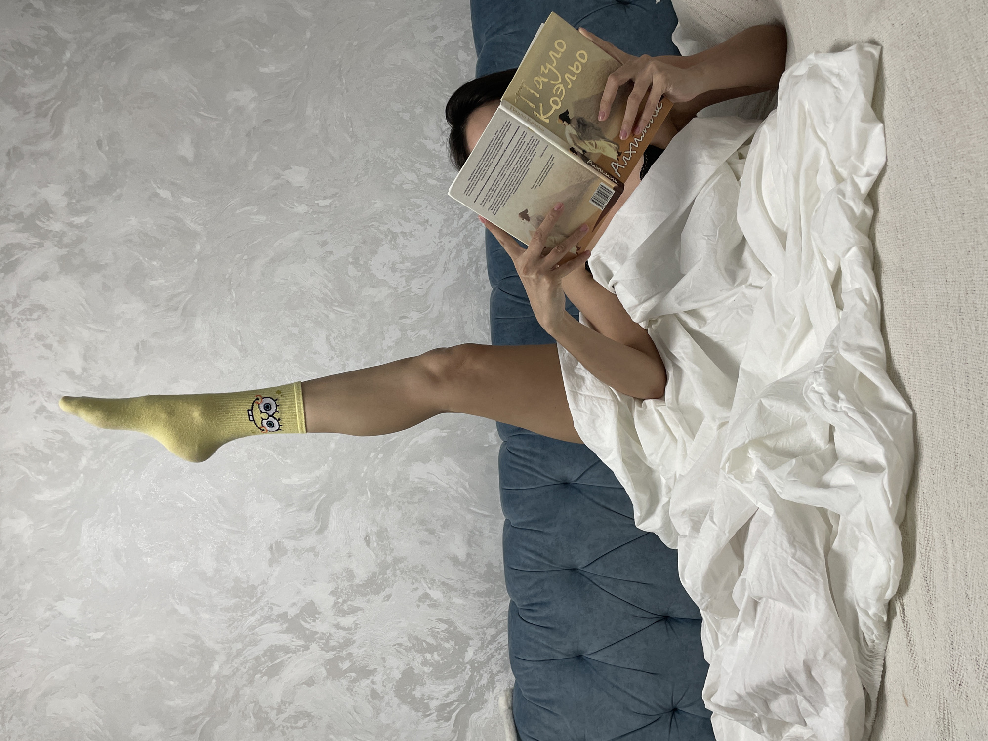 A person is reading a book in a bed. The person is lying on a blue tufted headboard, covered with white sheets. One leg, clad in a yellow sock with a cartoon character, is extended towards a textured white wall. The book being read has a light yellow cover with an illustration and text in Russian. The location is Izhevsk, Russia.