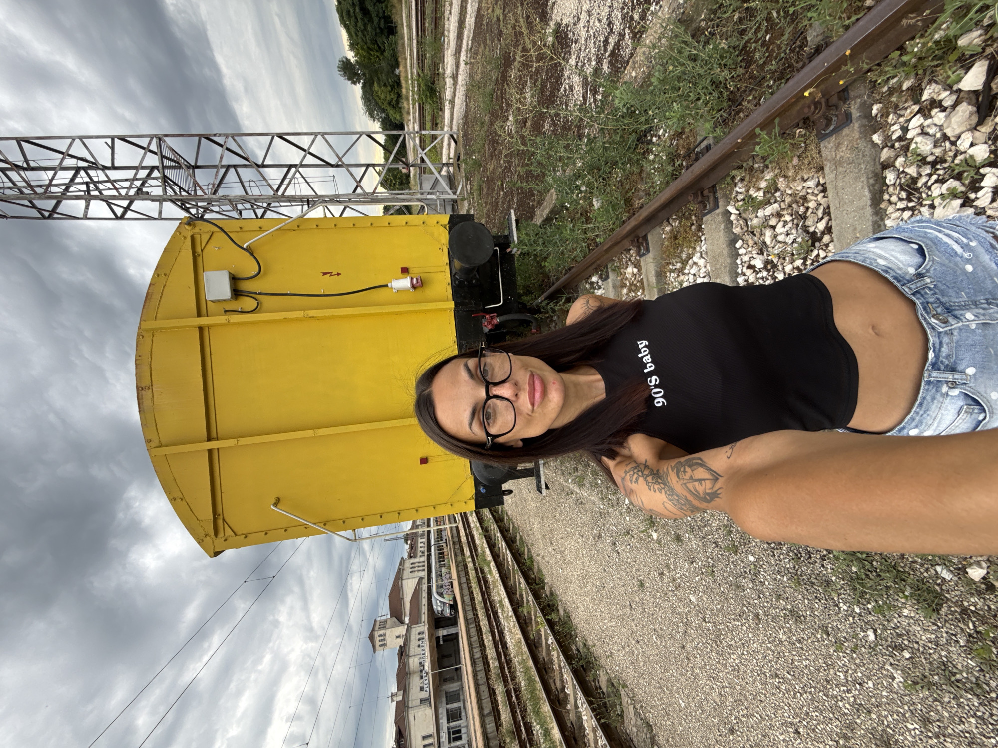 A woman with dark hair and glasses is taking a selfie at a train station in Pleven, Bulgaria. She is wearing a black crop top with "90's baby" printed on it and denim shorts. She has tattoos on her arm. Behind her, a large yellow train car is parked on the tracks. In the background, a train station building with a tower can be seen. The sky is cloudy, and there are trees and vegetation around the train tracks.