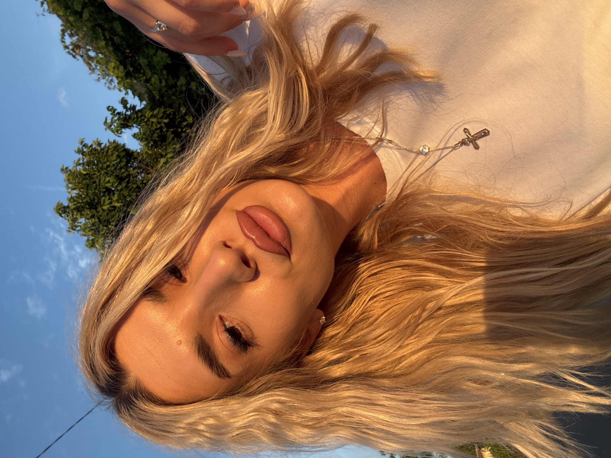 A young woman with blonde, wavy hair is captured in a selfie against a backdrop of a blue sky and lush green trees. The warm, golden light of sunset illuminates her face, highlighting her features and casting soft shadows. She is wearing a white t-shirt and a silver necklace with a cross pendant. A silver ring with a small gem is visible on her right hand, which is resting near her face. The setting appears to be outdoors, possibly in a park or garden area within the city of Skrat, Bulgaria, given the presence of trees and a clear sky suggesting daytime or early evening hours.