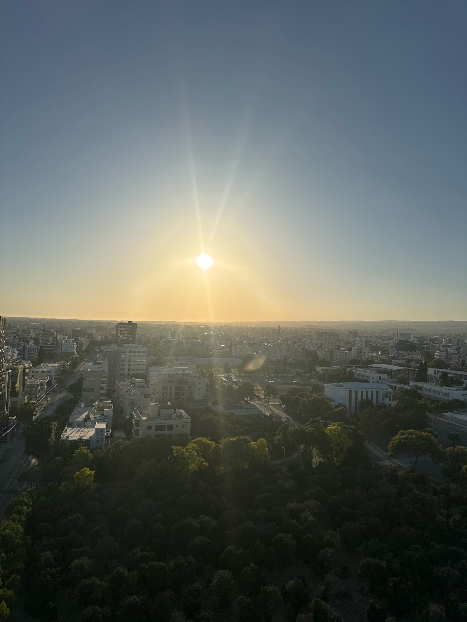 An aerial view captures a cityscape at sunset, characterized by a clear sky with the sun prominently positioned in the upper center, casting bright rays. The horizon displays a faint haze, suggesting a coastal city. In the foreground, dense, dark green foliage dominates, with some trees exhibiting yellowing leaves, indicative of autumn. Beyond the trees, a mix of urban structures is visible. The buildings are predominantly light-colored, ranging from white to beige, with some featuring darker architectural elements. Roads and sidewalks are discernible, with hints of streetlights. The scene appears to be the city of Limassol, Cyprus, given the distinctive architecture and the presence of palm trees in the distance, consistent with the Mediterranean environment. No individuals or specific actions are identifiable.