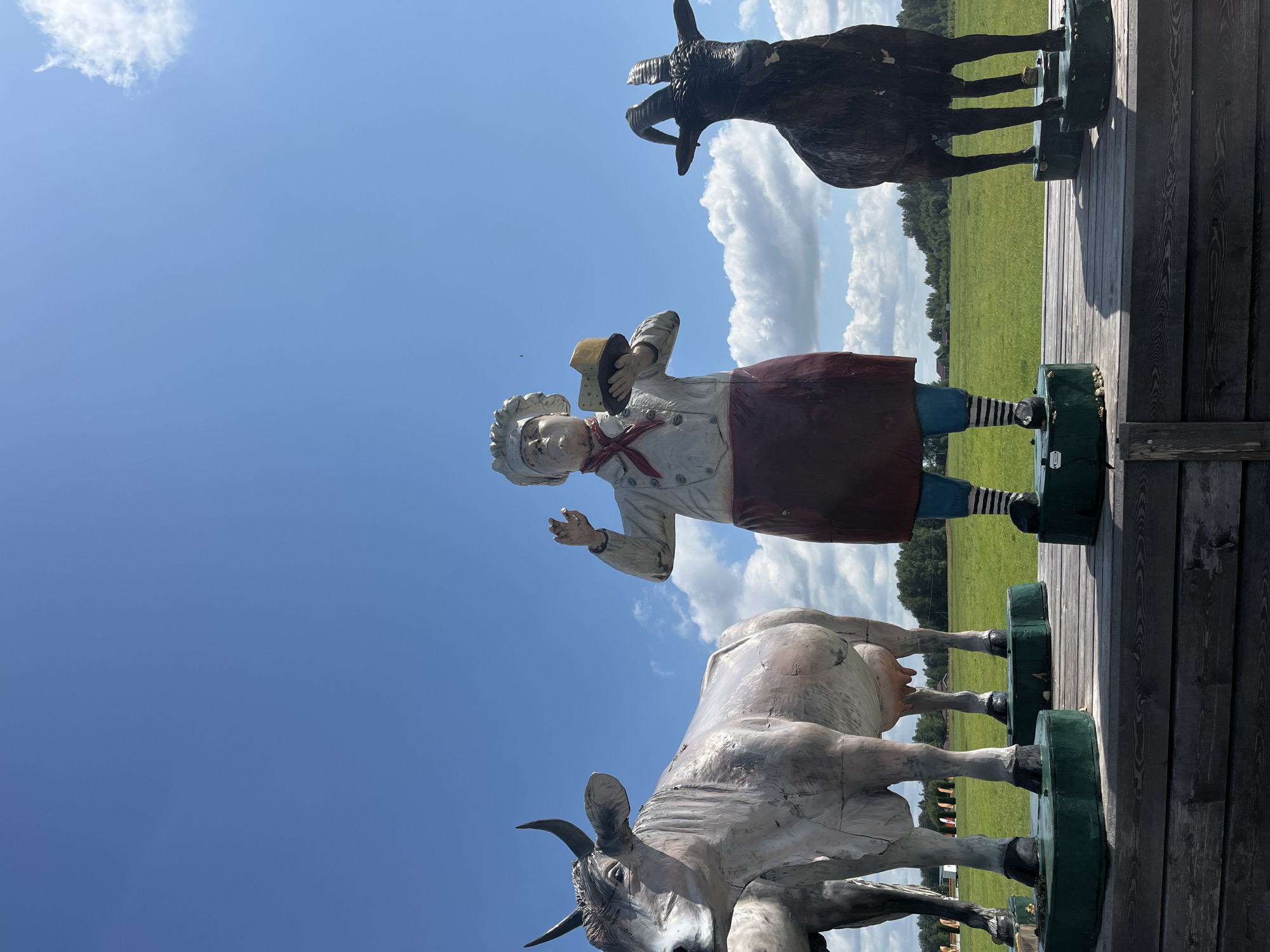 The image depicts a whimsical outdoor scene featuring life-size statues of a cow, a chef holding a cheese, and a goat. These statues are arranged on a wooden platform in a grassy field under a bright blue sky with scattered white clouds. The cow, positioned on the left, is a light gray color with large horns. The chef, in the center, is dressed in a white chef's coat, a red scarf, and a dark red apron, holding a block of cheese. To the right, the goat is dark brown with prominent horns. In the background, a line of trees forms the horizon, and a few small buildings are visible in the distance. The overall impression is that of an attraction or a themed exhibit, possibly related to dairy farming or a local festival, located in the city of Akhlebaikha (Kovriginskii S/S), Russia.