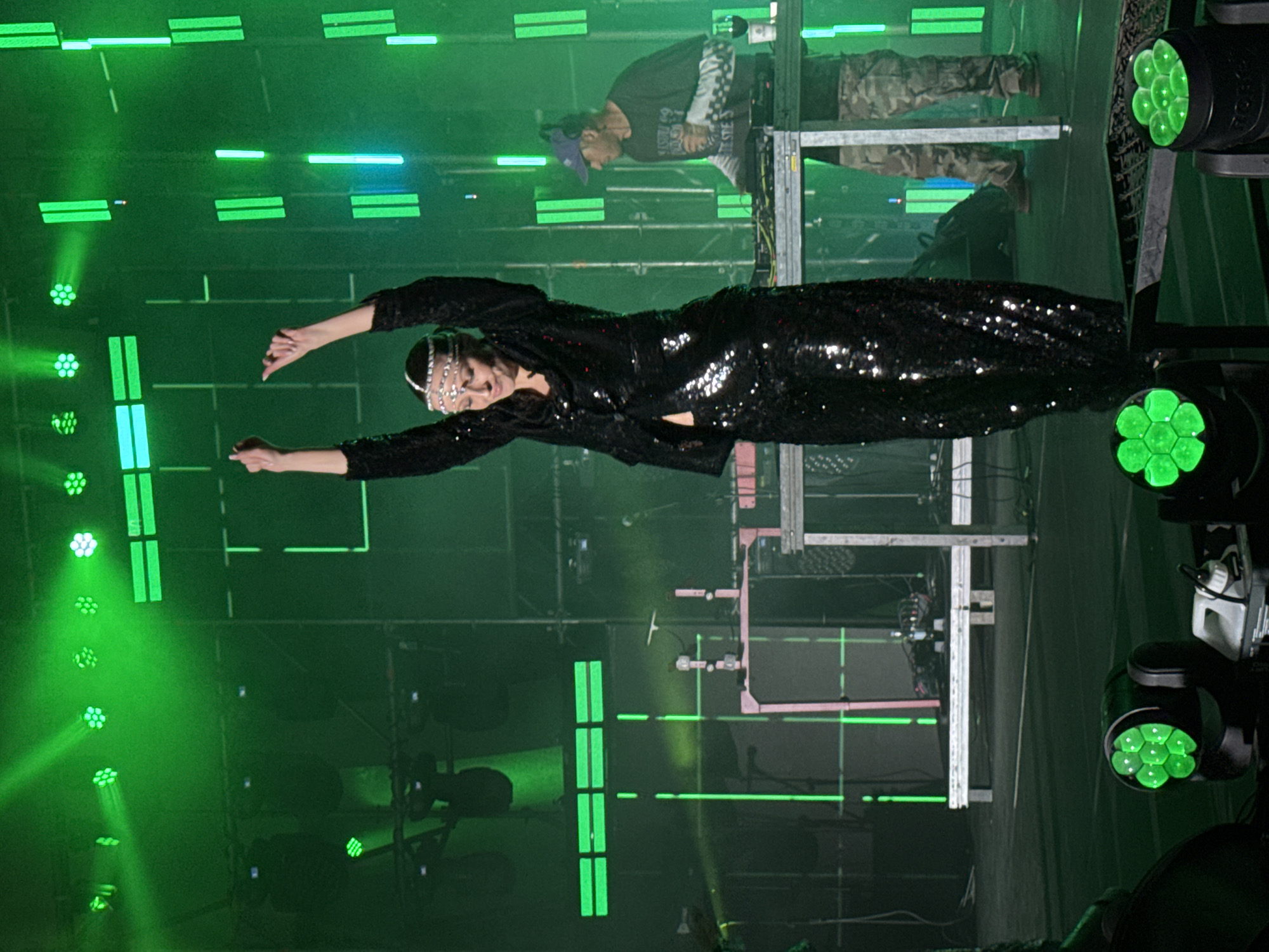 The image depicts a woman in a black sequined dress with a silver headpiece, standing on a stage bathed in green light. Her arms are outstretched. In the background, a person stands behind a DJ setup. The scene is a performance setting, likely a concert or show. The image was taken in Yerevan, Armenia.