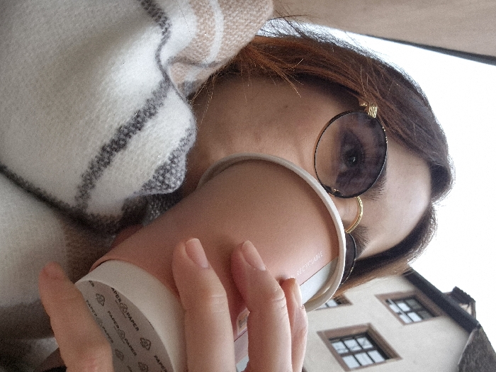 A low-angle close-up depicts a person holding a pink paper cup near their face, partially obscuring it. The individual wears round sunglasses with a gold frame, and a gold hoop earring is visible. They are wrapped in a white blanket with tan and gray stripes. Behind the person is a building with white walls and several square windows. The overall lighting appears soft and diffused. The cup has “PAPER” printed repeatedly on its rim.