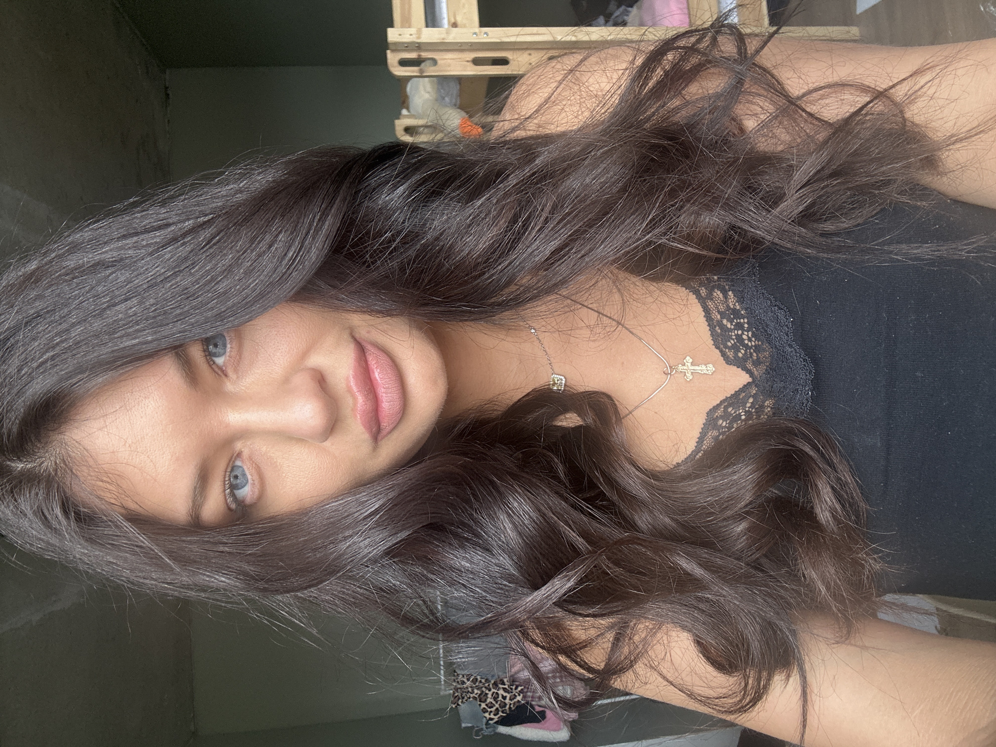 The image, rotated 90 degrees clockwise, captures a young woman from a medium-close perspective. Her head is on the left side of the frame, and she gazes directly at the viewer with striking light blue eyes. She has long, dark brown, wavy hair that cascades over her left shoulder and chest. Her lips are slightly parted, revealing a soft, neutral expression, and she appears to be wearing a nude-pink lip color.
She is dressed in a black tank top featuring delicate black lace trim along the neckline. A thin silver necklace adorns her neck, displaying a small cross pendant and another small, possibly square-shaped charm.
The background is an indoor setting, appearing dimly lit and somewhat indistinct. A light-colored wooden structure, possibly shelving or part of a bed frame, is visible in the upper right quadrant. Further details in the background include hints of a leopard-print fabric and what appears to be a pink and white shoe, partially covered by her hair in the lower right. The lighting is soft, primarily illuminating her face and hair. There is no visible text in the image. The scene suggests a casual, personal moment indoors in Khimki, Russia.