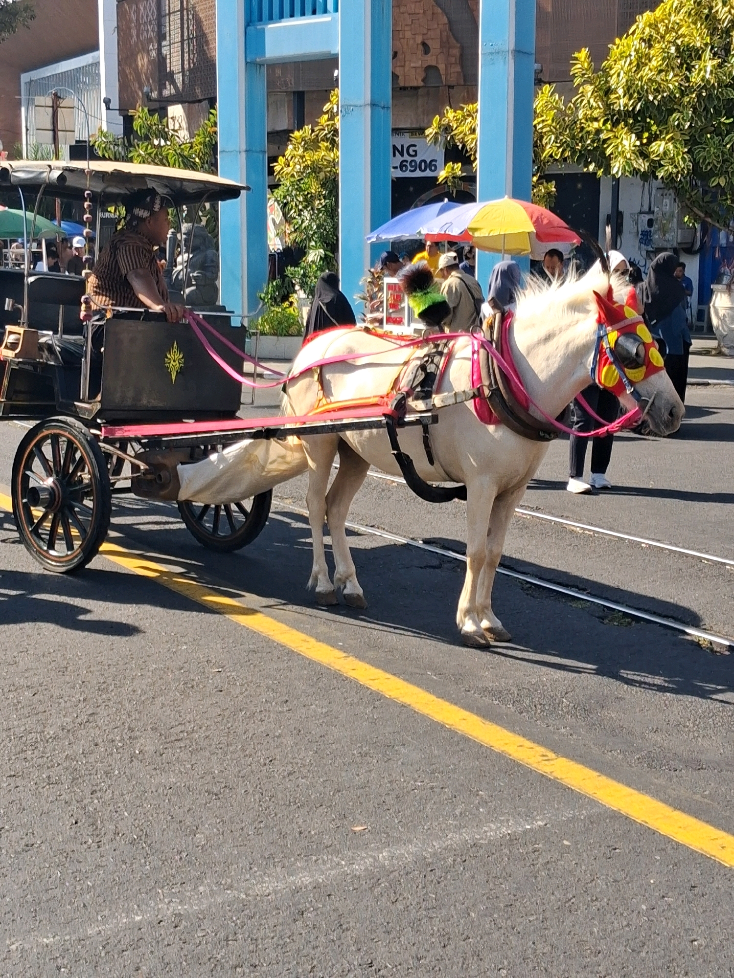 A horse-drawn carriage, known locally as a "andong," is on a paved street in Surakarta, Indonesia. The carriage has two large spoked wheels in the front and two smaller ones in the rear, all black with silver accents. A brown, wooden seat for the driver is positioned at the front of the carriage, with a man wearing a striped shirt and a patterned head covering driving. Pink straps extend from the carriage to the horse. The horse is white with a decorated bridle featuring red and yellow accents around its eyes and forehead. The horse is equipped with a saddle and harnesses. In the background, a blue architectural structure with a weathered, brown tiled facade is visible. Several people are present on the street, including a woman in a black hijab and a man in a blue shirt. The street is marked with a yellow line. The overall scene appears to be a public area, likely a tourist attraction.
