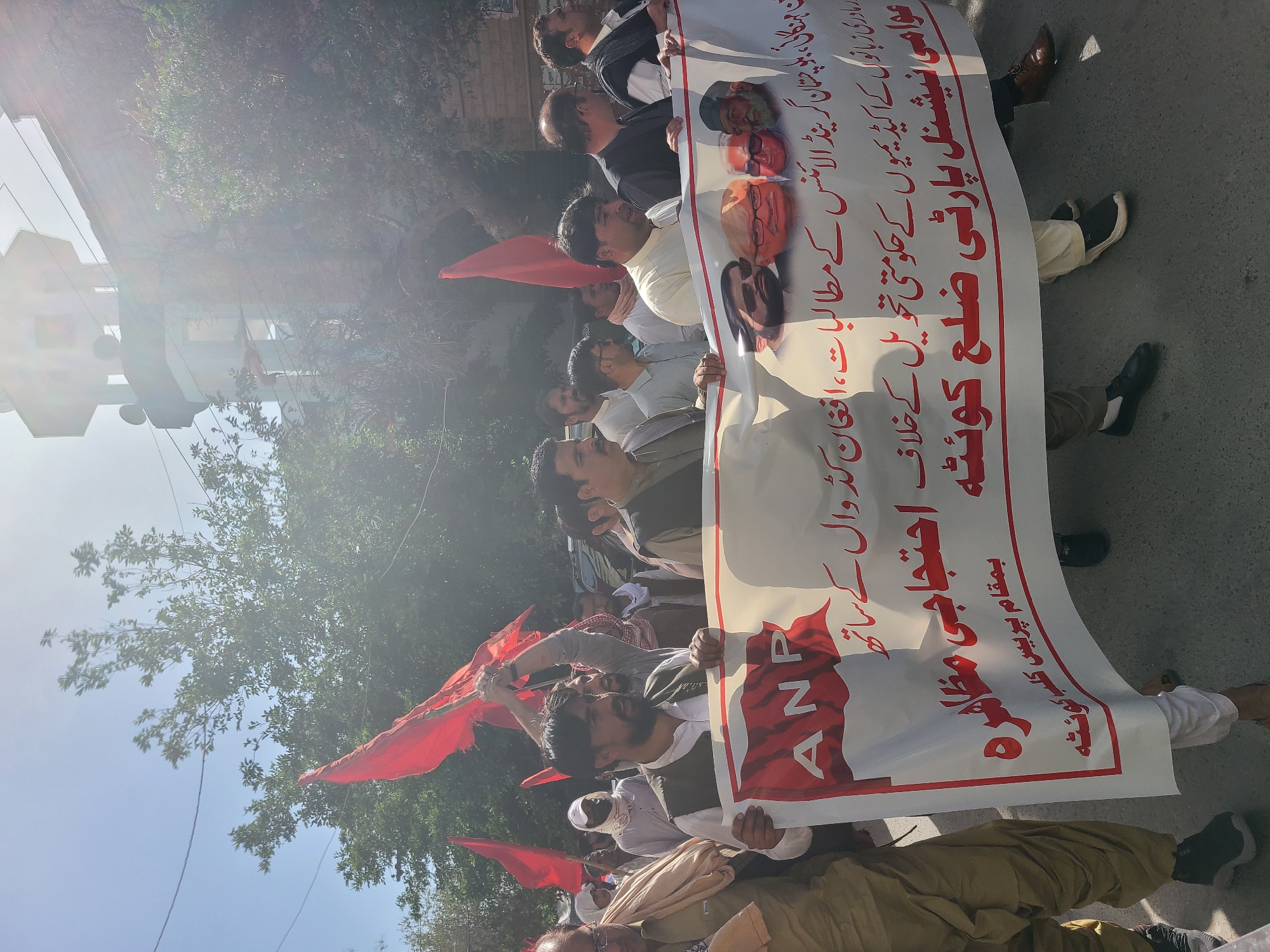 A group of people are marching in Quetta City Tehsil, Pakistan. They are holding a banner with text written in Urdu that reads "Awami National Party District Quetta" and mentions "protest against government control of mother tongue academies." The banner also shows portraits of individuals and the initials "ANP" in red. Some participants are holding red flags. The march is taking place on a street in daylight.