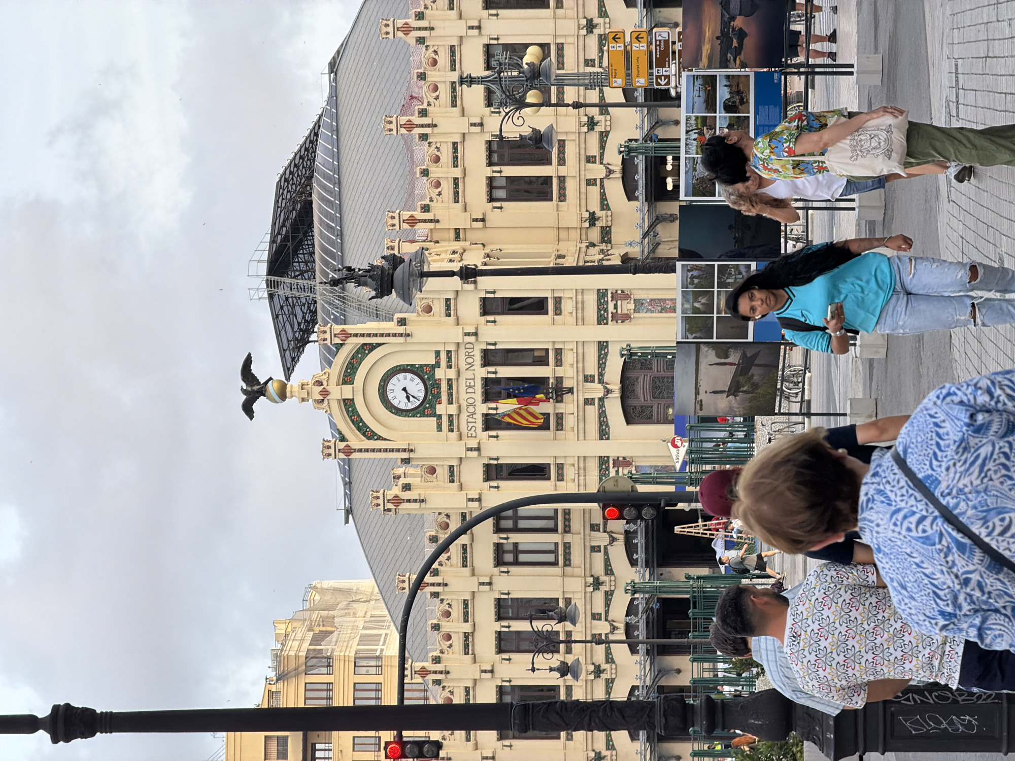 In an outdoor urban setting, several people are walking on a paved walkway in front of a large, ornate building. The building, identified by a sign as "ESTACIÓ DEL NORD," appears to be a train station, featuring a clock on its facade and flags displayed prominently. An eagle sculpture perches atop a decorative element on the building's roof.
In the foreground, a group of individuals is captured from behind, their backs to the camera, as they move away. One person in this group wears a white t-shirt with a colorful floral pattern, while another is dressed in a blue and white patterned shirt.
Further into the scene, a woman in a bright turquoise t-shirt and ripped denim jeans walks towards the camera, holding a smartphone in her left hand. To her right, another woman, wearing a white top with a colorful floral print and green pants, carries a canvas tote bag.
Several large framed photographs are displayed on stands to the left of the main building, showcasing various landscapes and water scenes. Streetlights and traffic signals are also visible throughout the scene. The sky is overcast, suggesting a cloudy day.
