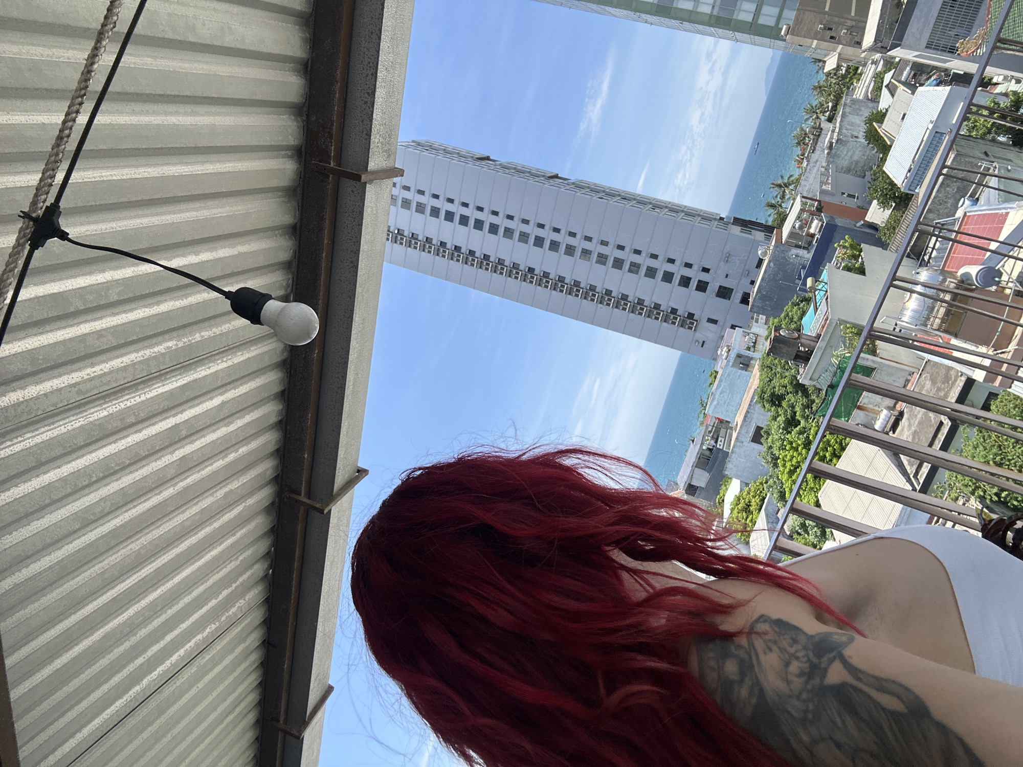 An outdoor shot captures a woman with vibrant red hair, facing away from the camera, on a balcony with a view of the city and ocean in Đà Nẵng, Vietnam. The woman is wearing a white top, and a portion of a tattoo is visible on her left arm. The balcony is covered by a corrugated metal roof, and a bare light bulb hangs from a cord above. In the background, several buildings, including a tall high-rise apartment building, are visible, as well as the ocean and a clear blue sky with scattered clouds. The overall scene suggests a sunny day with a tropical atmosphere.