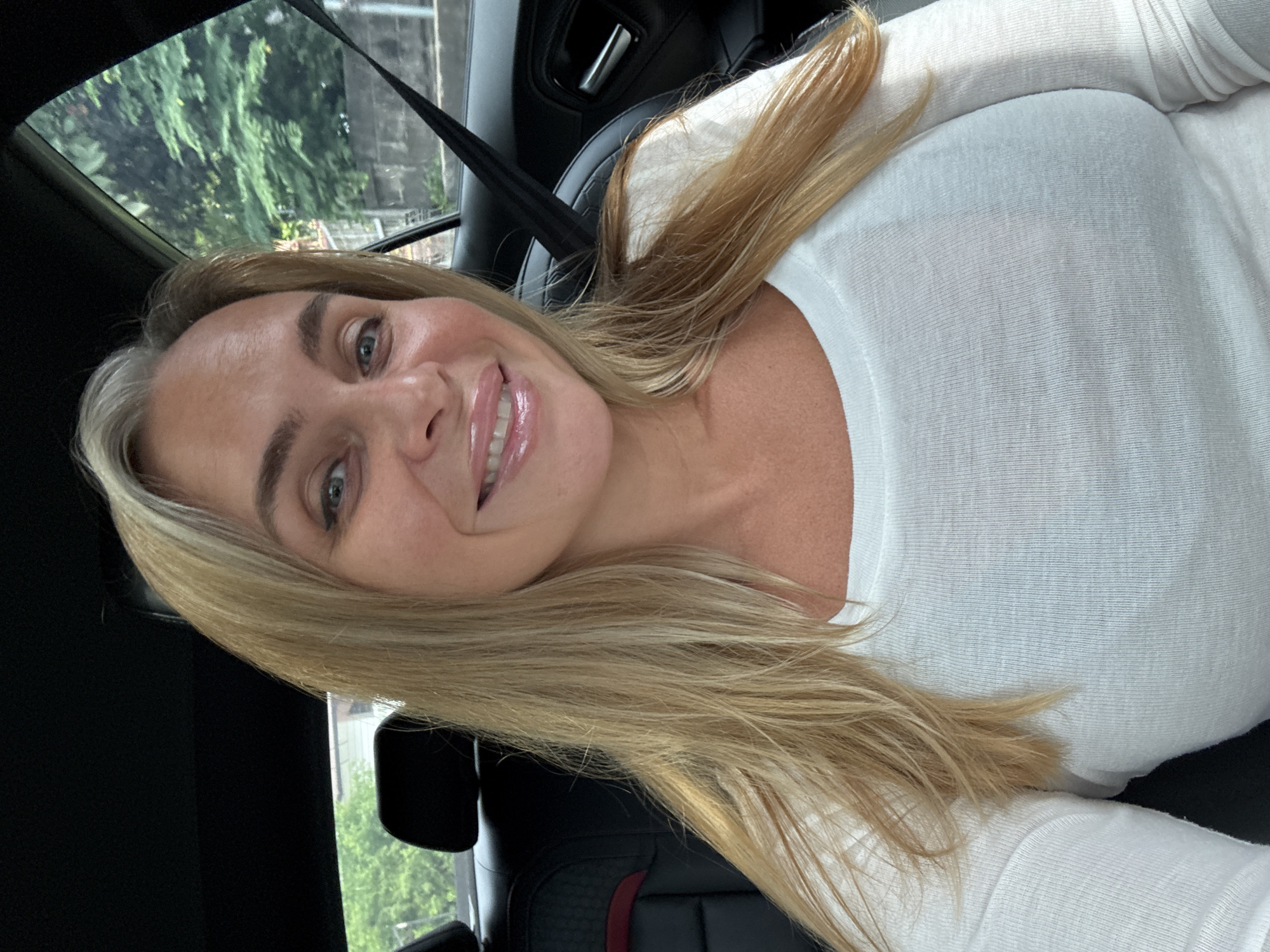 A blonde woman with blue eyes and full lips is seated in the back seat of a car, likely a luxury model given the leather interior and accent stitching. She is wearing a white long-sleeved top. The interior of the car is dark, with black leather seats and headrests visible. A black seatbelt extends across her right shoulder. Through the car window, lush green trees and a stone wall are visible, suggesting an outdoor environment. The background appears to be a street or residential area, potentially within a city, with some hints of buildings. The lighting suggests it is daytime, possibly overcast or late afternoon, as there are no harsh shadows. The image is a selfie, with the camera positioned at a slight upward angle. The woman is smiling and looking directly at the camera. The car's interior is clean and well-maintained. No other individuals are visible in the frame. The overall impression is a casual, personal photograph taken during a car journey.