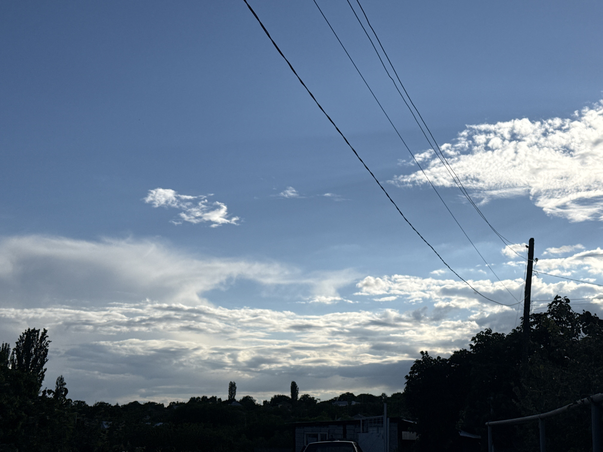 The image depicts a sky filled with scattered clouds. Power lines stretch diagonally across the frame from the upper left to the lower right. In the foreground, a silhouette of trees and a utility pole is visible against the brighter sky. The lower portion of the image shows the dark silhouettes of rooftops and vegetation. The scene appears to be an outdoor environment in a city.