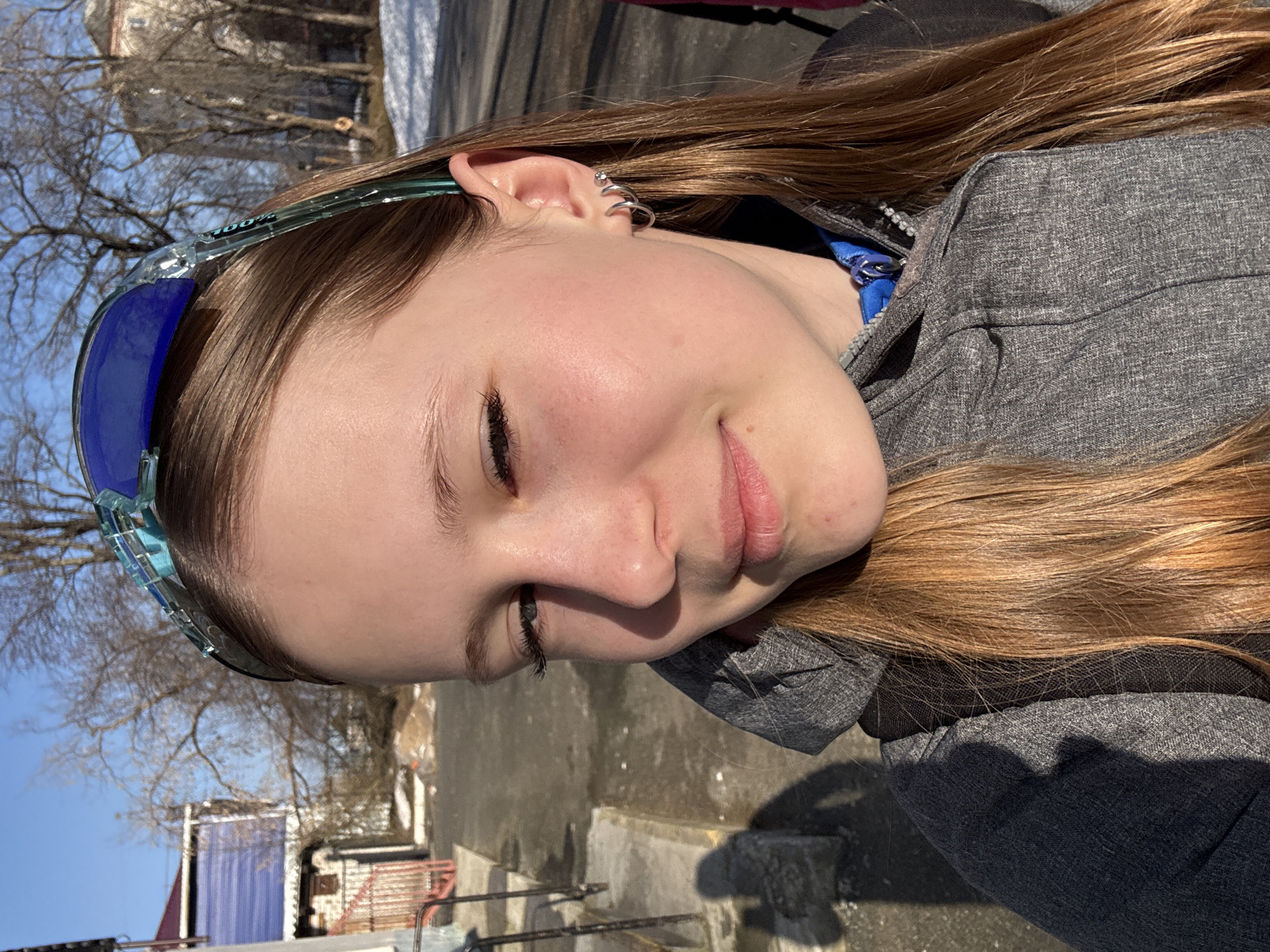 A close-up, angled portrait shows a young woman with long, light brown hair. She is wearing sunglasses with blue lenses on her head. Her left ear is visible, adorned with multiple silver earrings. She wears a grey zip-up hoodie with a blue drawstring.
The background is a cityscape, featuring bare trees, buildings with snow on top, and pavement. Based on visual landmarks and available information, the location appears to be Berezniki, Russia.