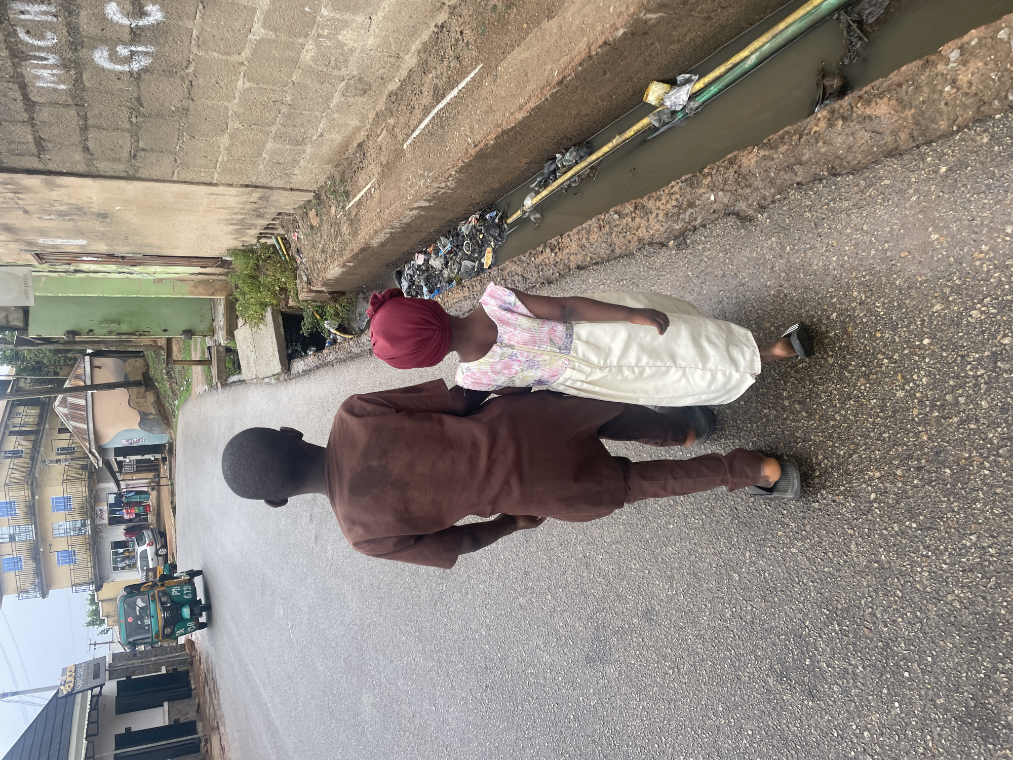 Two individuals, one wearing a brown, long-sleeved garment and dark shoes, and the other wearing a red head covering, a light-colored, floral-patterned top, and a long, white skirt, are walking on a paved road in Jos, Nigeria. A drainage ditch filled with trash and pipes runs alongside the road. Buildings and a vehicle are visible in the background. A building with the number 55 written on it is also partially visible.