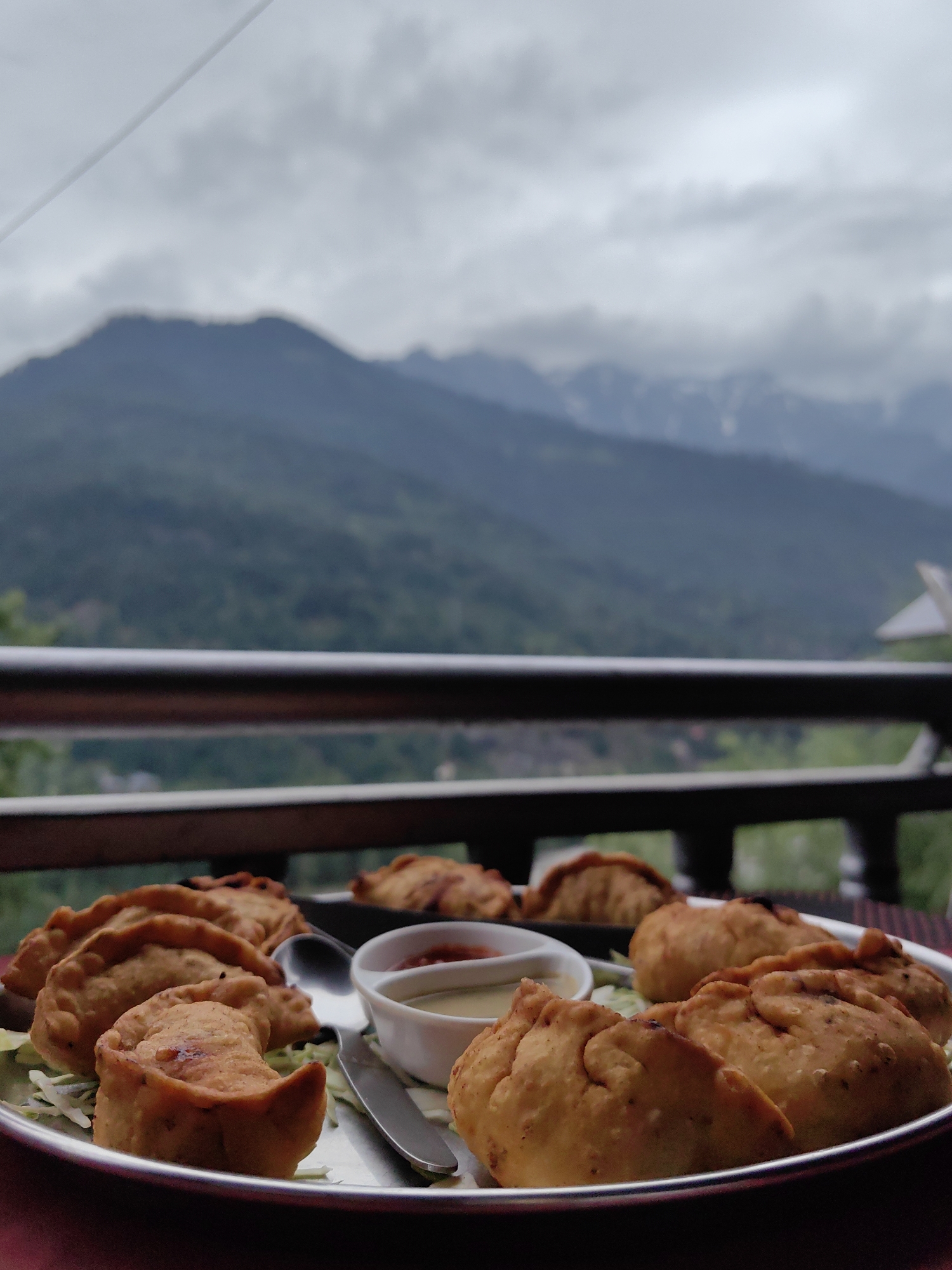 A plate of fried dumplings sits on a table in front of a mountain landscape in Vashisht, India. The dumplings are golden brown and appear to be filled with a savory mixture. They are served with two dipping sauces, one red and one yellow, in small white bowls. A spoon and a fork are also present on the plate. In the background, lush green mountains rise towards a cloudy sky. The overall scene suggests a delicious meal enjoyed with a breathtaking view.