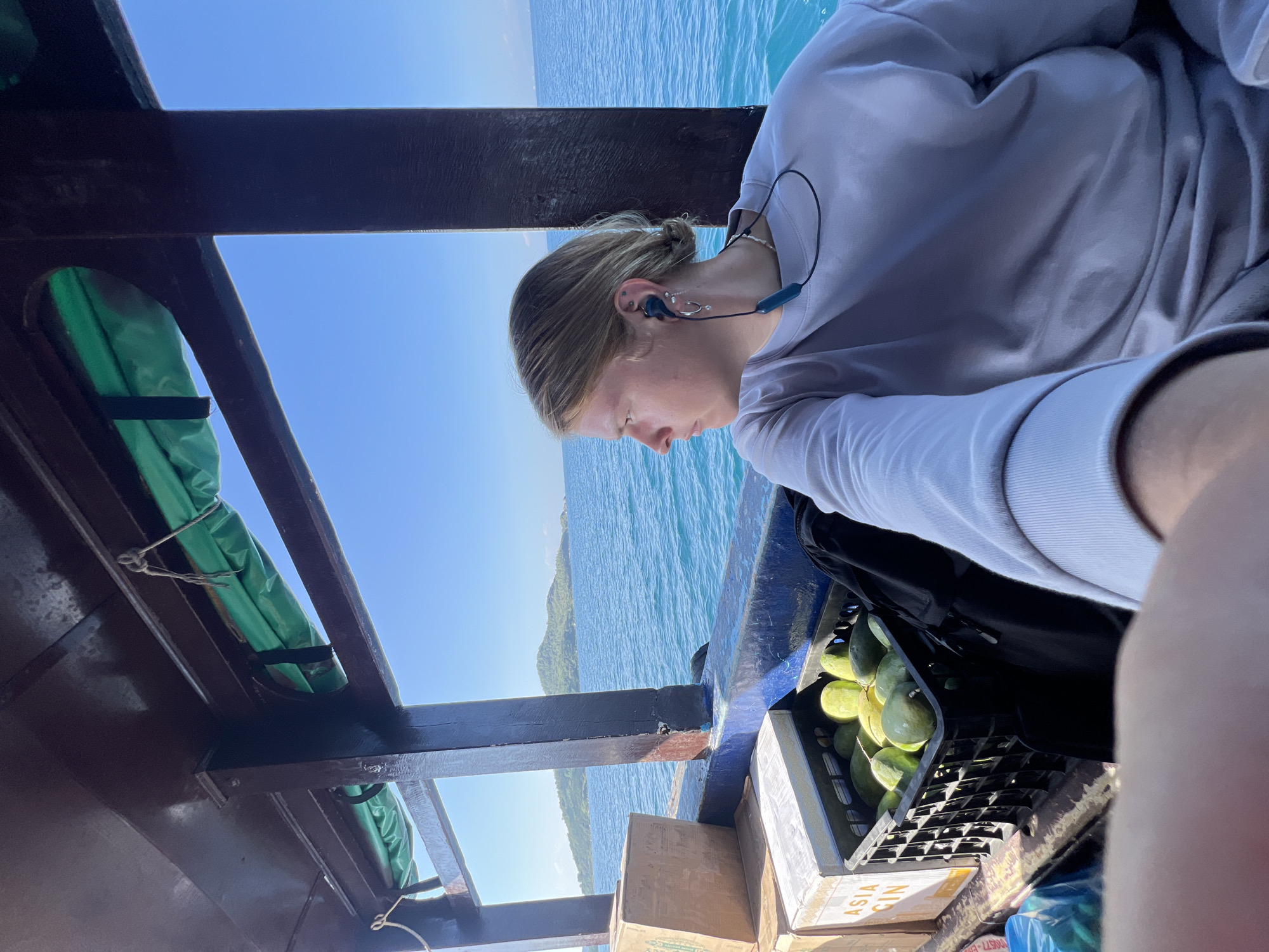 A person with fair skin and light blonde hair is on a boat, possibly a ferry, on the coast of Bac Nha Trang Ward, Vietnam. The person is wearing a light gray long-sleeved shirt and blue earphones. They are looking down, possibly at the water. A black crate filled with green mangoes sits next to them, resting on top of cardboard boxes labeled "Asia Gin." The boat has a wooden frame structure with green life vests secured to the ceiling. The ocean is visible in the background, along with a hazy blue sky and a distant, green mountainous coastline.