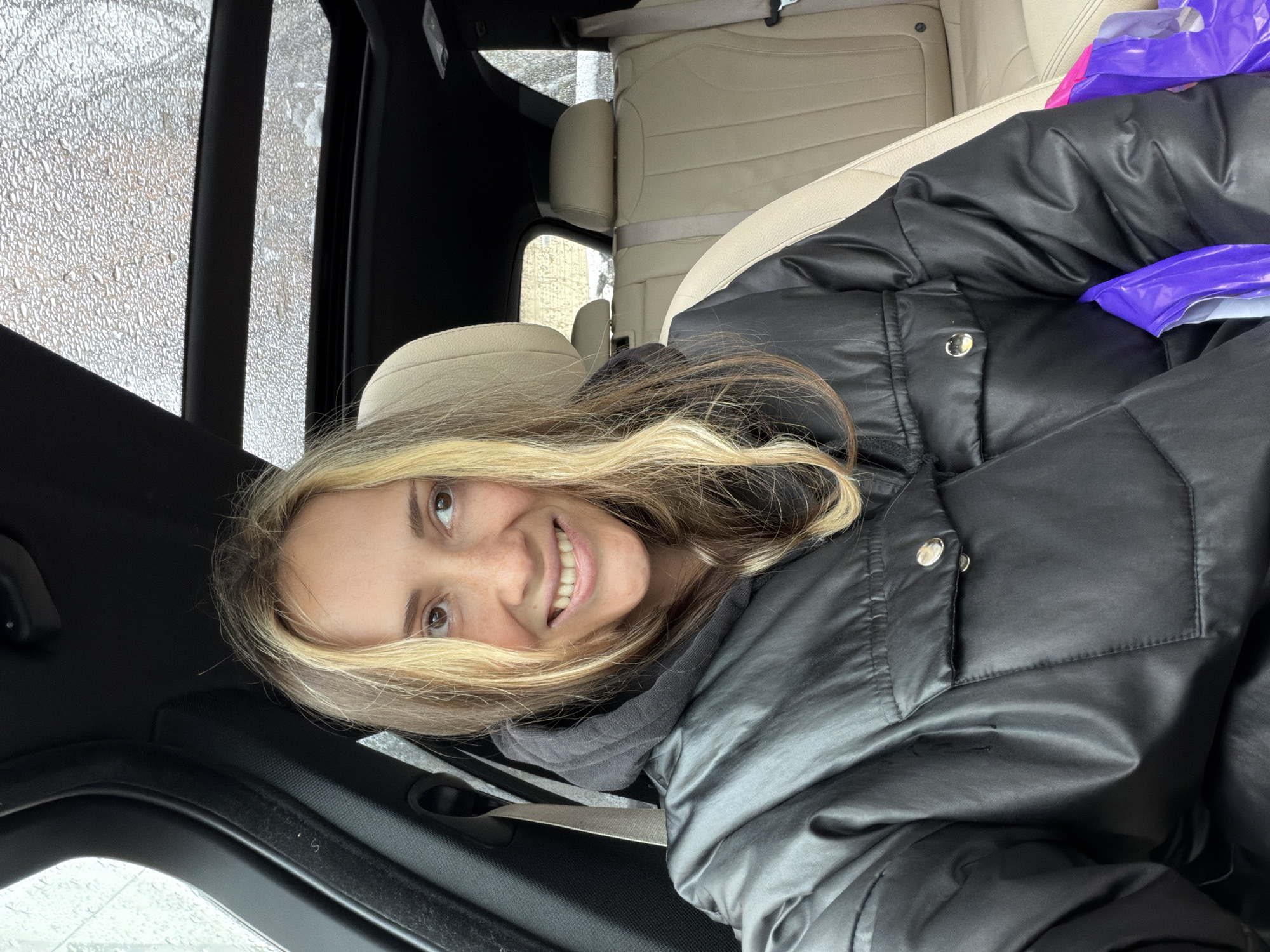 A young woman with long, blonde and light brown hair smiles widely at the camera, revealing her teeth, while seated inside a car in Khimki, Russia. Her brown eyes are open and engaging. She is wearing a black puffer jacket over a dark grey hooded top, with a silver button visible on her jacket. A bright purple plastic bag is partially visible on the seat next to her.
The interior of the car features light-colored (beige or cream) leather seats and dark trim. Through the windows, numerous water droplets and streaks are visible, indicating rain or sleet outside. This suggests a wet and potentially cold weather day. The woman appears happy and relaxed, posing casually for the photograph while inside the vehicle, likely for warmth or shelter from the weather. No discernible text is visible.