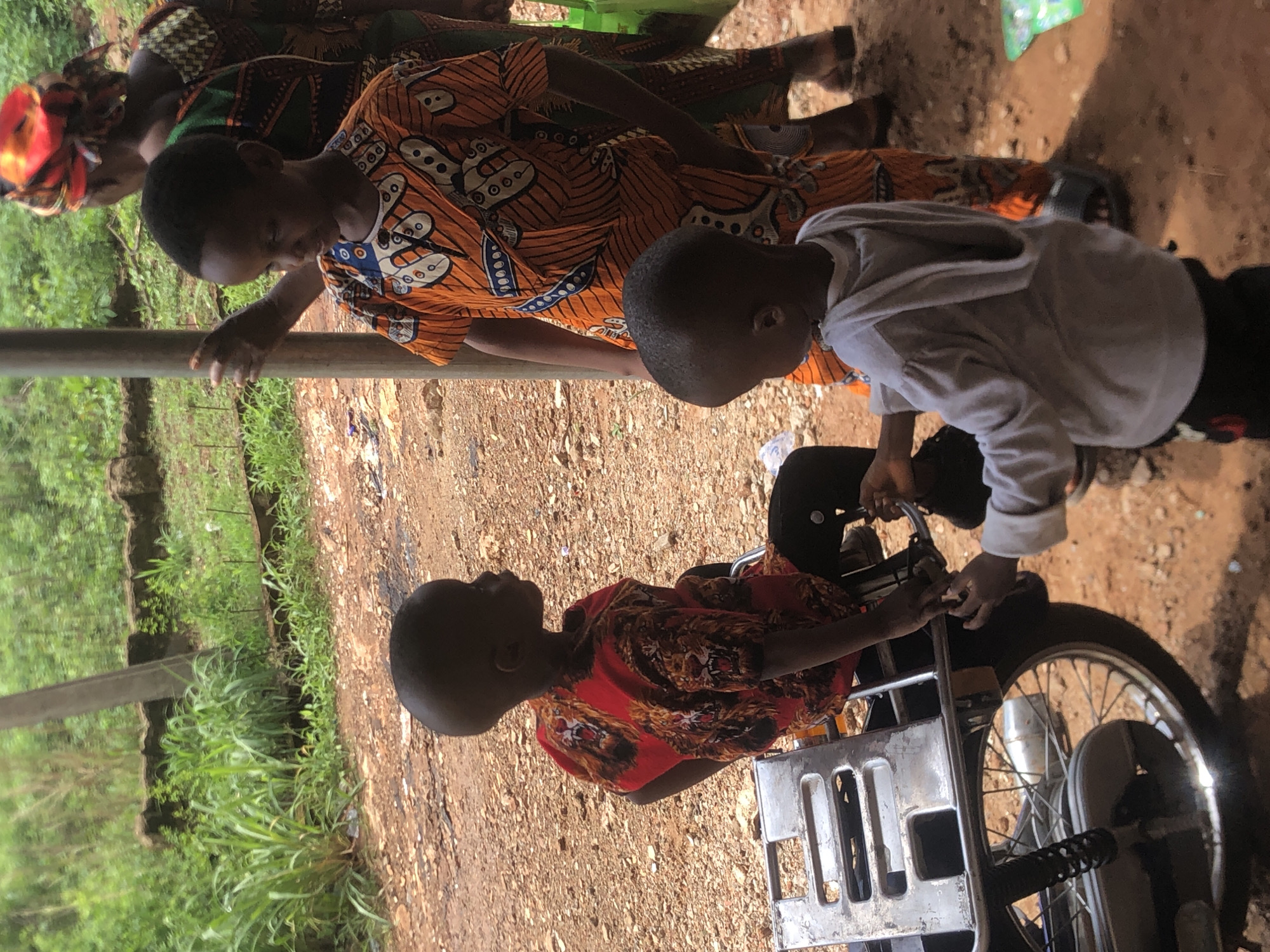 Three children stand on a bare dirt surface, likely soil. A motorized vehicle, possibly a motorcycle, is present, with one child seated. A second child, clad in a grey jacket, appears to be interacting with the motorcycle's controls. The third child, along with an adult wearing a head wrap, stands nearby, possibly observing. The environment includes green vegetation to one side and what looks like the support structure of a building above the people. The location is Nkwelle, Nigeria.