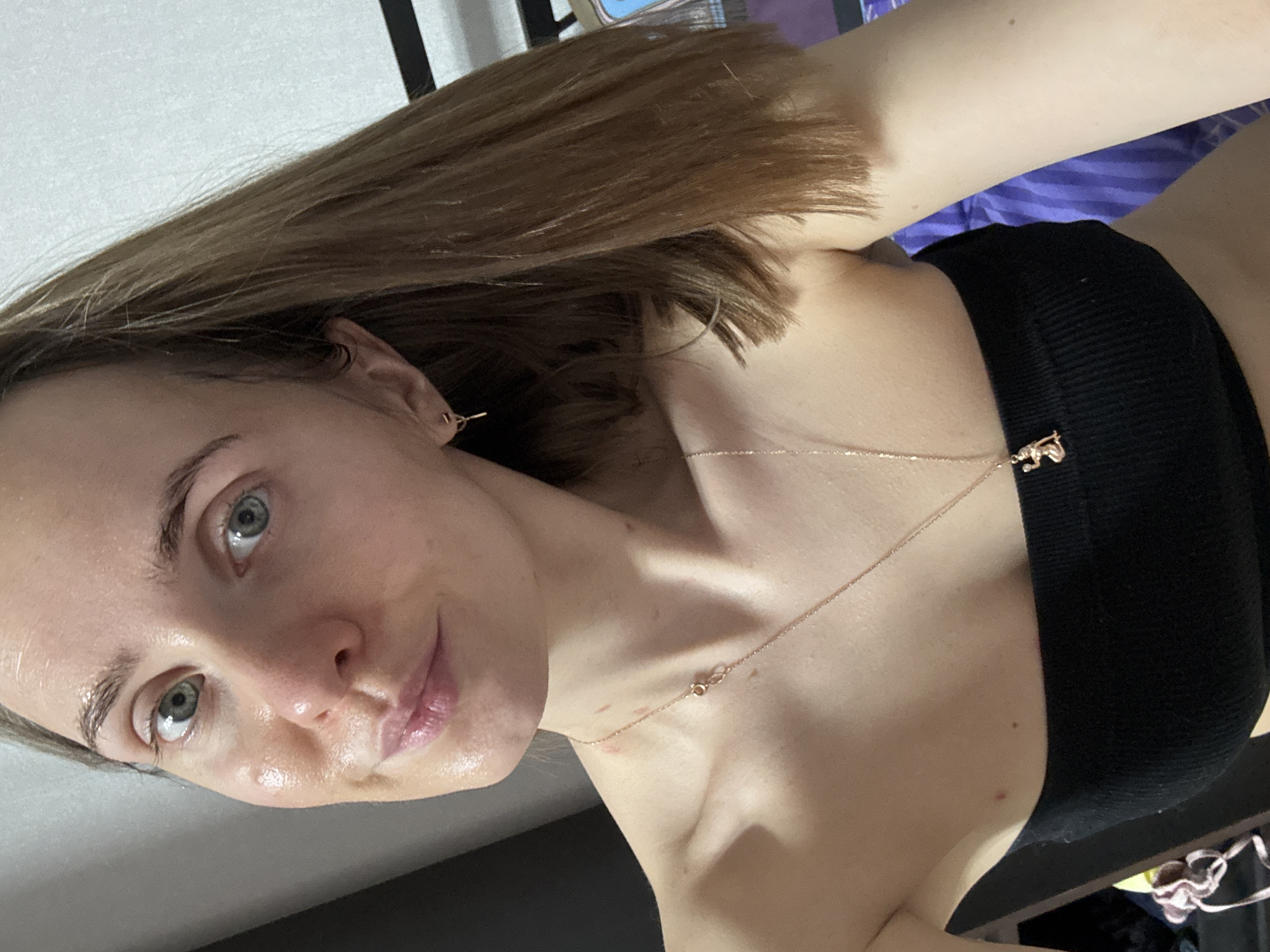 A selfie of a woman with shoulder-length brown hair, light skin, and blue eyes. She is wearing a black top and a delicate gold necklace with a small pendant. The background appears to be an interior space with blurred objects, possibly a room. It is impossible to determine if the location is Perm, Russia without further information.