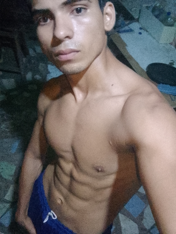 The image portrays a young adult male, light-skinned with dark hair and eyes, captured indoors. The scene is situated in Juan Griego, Venezuela. The male subject is shirtless, exhibiting a muscular physique with visible abdominal definition and developed shoulder musculature. He is wearing blue athletic shorts with a white drawstring. The subject faces the camera directly, holding a neutral expression. His torso is slightly angled, with his right arm partially visible, bent at the elbow. The background is dimly lit and largely out of focus. A light blue-gray surface is discernible behind the subject's left shoulder (viewer's right), possibly a wall. To the subject's right (viewer's left), the background is darker and indistinct. The floor in the lower right section of the image displays a patterned surface, possibly blue and dark-toned tiles. Illumination appears to be from a direct, localized source, casting shadows that emphasize the subject's musculature. The image possesses a grainy quality.