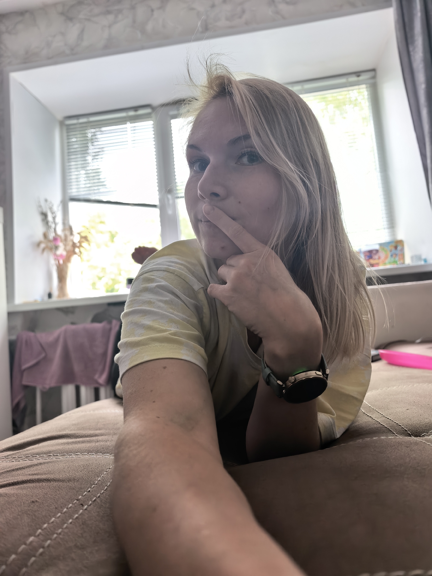 The image captures a young woman in a close-up, low-angle shot, seemingly taken as a selfie. She has blonde hair and light-colored eyes, looking directly towards the camera with a thoughtful or slightly playful expression. Her right index finger is lightly touching her nose or upper lip. She is wearing a light yellow tie-dye t-shirt, and a smartwatch with a dark band, rose gold casing, and a glowing green sensor is visible on her left wrist.
The setting is indoors, likely a home environment. The woman is lying on a brown upholstered surface, which appears to be a couch or bed. In the background, a large window with white horizontal blinds is visible, allowing natural daylight to stream in. Outside the window, green foliage suggests a daytime scene with pleasant weather. On the windowsill to the left, a vase containing dried grasses and pink artificial flowers is placed. A dark grey curtain partially frames the right side of the window. Another small, colorful item, possibly a book or decorative box, is on the windowsill to the right. A pink object, resembling a brush or remote, lies on the couch near her arm. The walls have a light, possibly textured or marbled finish. There is no visible text in the image. The scene takes place during the day, in Sterlitamak, Russia.