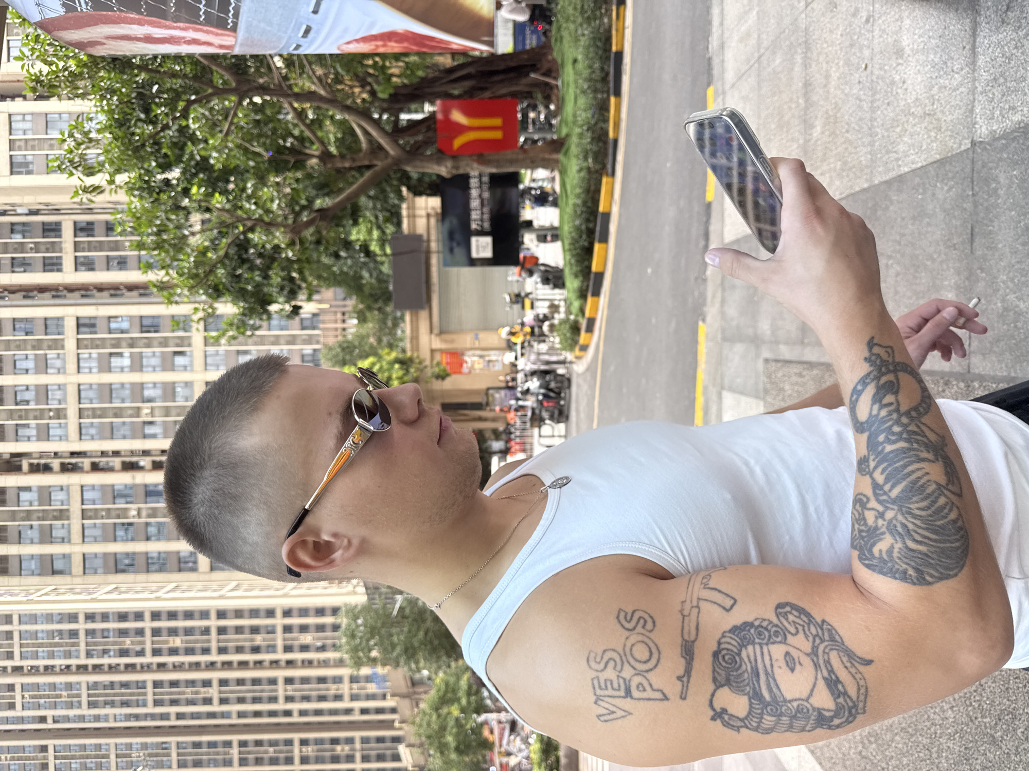 A young man with a buzz cut and sunglasses is standing on a sidewalk in Haizhu District, China, looking at his phone. He is wearing a white tank top and has several tattoos on his left arm, including text that appears to read "VES POS," an image of a rifle, a blindfolded figure with a snake, and a tiger. He is also holding a cigarette in his right hand. In the background, there are tall buildings, trees, and street signs, including a red sign with yellow logos that resembles a McDonald's arch. The street is paved and has yellow and black striped markings along the curb.