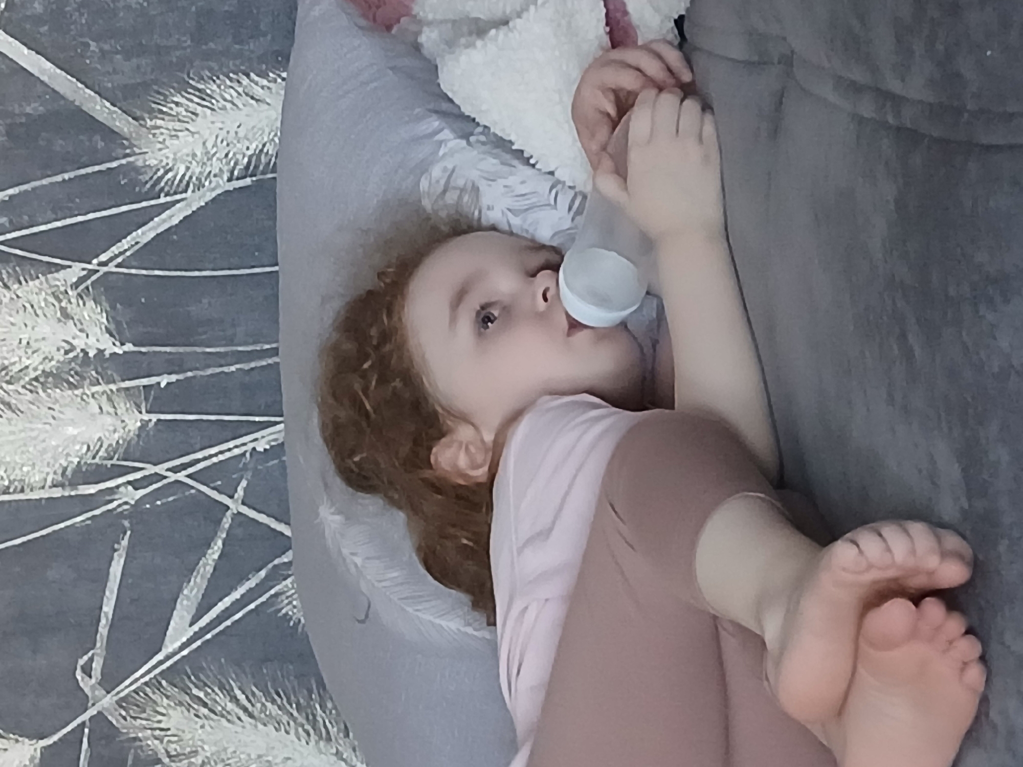 A young child with reddish-brown curly hair is lying down, possibly on a couch or bed, and drinking from a bottle. The child is wearing a light pink shirt and darker pink pants. The background includes a gray surface with white decorative patterns and a soft, possibly furry, white object. The scene appears to be indoors. Марковский is not identifiable in the image.