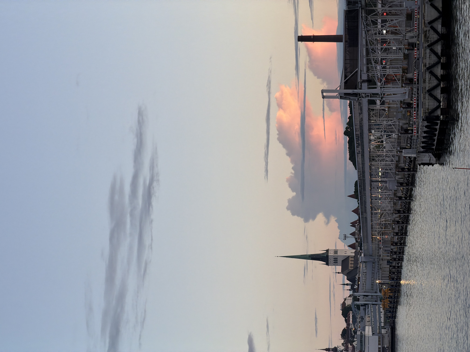 This image depicts a twilight scene at the Tallinn Port in Tallinn, Estonia. The foreground features a body of water, likely the Baltic Sea, reflecting the soft light of the setting sun. A long, modern pier stretches across the middle ground, equipped with jetties, walkways, and industrial structures. In the background, the historic skyline of Tallinn's Old Town is visible, dominated by the spires of churches, most notably the iconic St. Olaf's Church. The sky above is a canvas of pastel colors, with streaks of pink and orange from the sunset illuminating scattered clouds, interspersed with softer, greyer cloud formations. The overall atmosphere is serene and tranquil, capturing the transition from day to night over a bustling port city.