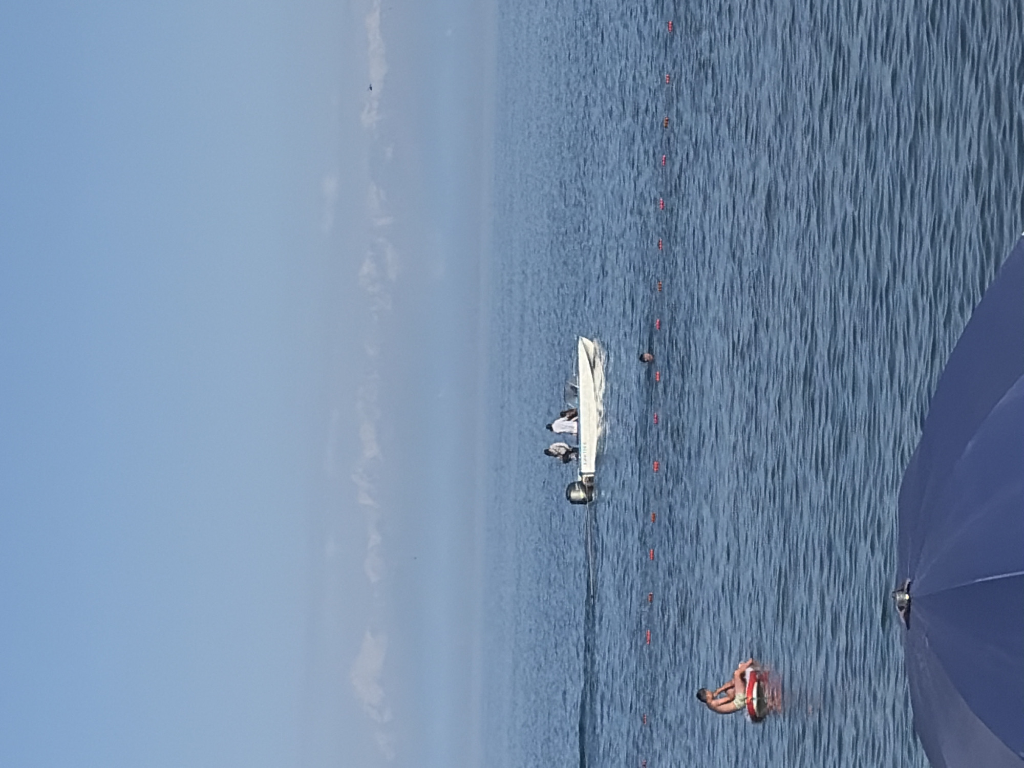 A wide shot of a calm body of water under a clear blue sky, with a hazy, mountainous horizon in the distance. In the foreground, the top of a dark blue umbrella is visible, suggesting it is on a beach. Further out, a line of red buoys floats on the surface, marking a boundary. A person is seated on a red and white inflatable raft to the left of the frame. In the middle ground, a white motorboat with at least three occupants is cruising. To the right, a person's head is visible above the water, swimming near the buoys. A very small, dark object, possibly a bird or drone, is visible in the sky above the horizon.
