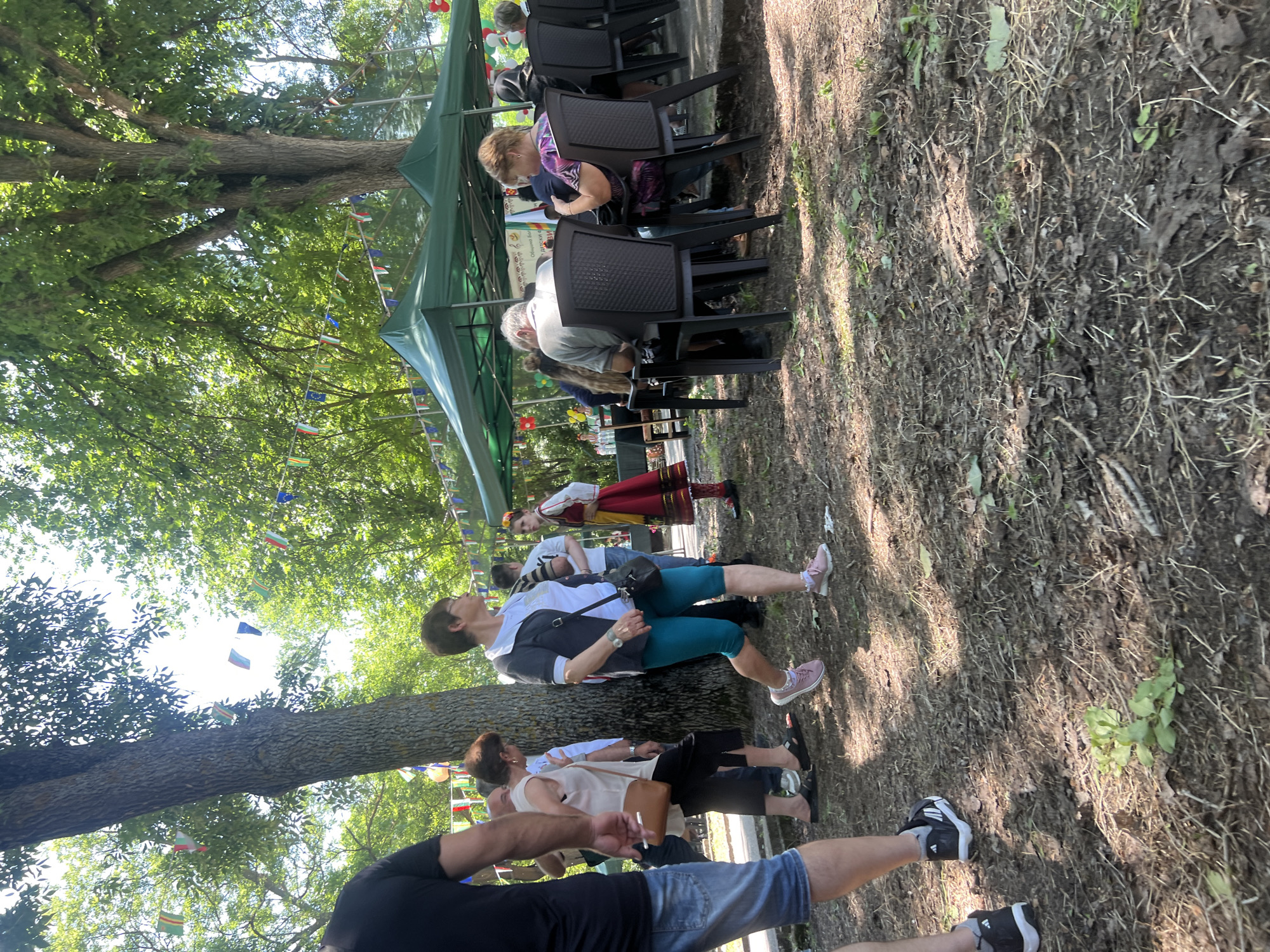This detailed description pertains to an outdoor gathering in Bolyarovo, Bulgaria.
The image captures an outdoor event under a canopy of large, leafy trees on a sunny day, with bright, dappled sunlight on the ground. The scene is populated by approximately 15-20 individuals engaged in various activities.
In the immediate foreground on the left, a man wearing a black t-shirt, blue denim shorts, and black athletic shoes is partially visible, his left arm and torso prominent. The ground is dry earth, uneven, and covered with sparse dry leaves, twigs, and small patches of green vegetation.
In the mid-ground, several individuals are walking from left to right. A woman with short brown hair, dressed in a white sleeveless top, black trousers, and carrying a tan shoulder bag, walks past a tree trunk. Alongside her, a woman with short, light brown hair and glasses wears a light t-shirt, a dark vest, teal capri pants, and pink athletic shoes, carrying a dark shoulder bag. Further right, a young woman or girl in traditional Bulgarian attire stands with her back partially to the camera, wearing a white top with red embroidery, a red and yellow skirt, a red headband, and dark shoes.
On the right side of the image, two dark green pop-up canopy tents are erected, providing shade. Beneath these tents, several people are seated on dark plastic chairs around tables. One man, seen from the back, has short gray hair and wears a light gray short-sleeved shirt. Next to him, a woman, also seen from the back, has brown hair and wears a purple patterned top. A white banner visible under the right tent displays Cyrillic text, partially legible as "Община Болярово" (Municipality of Bolyarovo) and "Детски Празник" (Children's Festival), confirming the event's nature and location.
Strings of colorful flags and bunting, including Bulgarian flags (white, green, red stripes) and European Union flags (blue with yellow stars), are suspended overhead between the trees and poles, stretching across the upper and middle portions of the scene. A cluster of red, white, and green balloons is visible in the background near the right tent. The setting appears to be a park or wooded area, with significant tree cover creating a shaded, convivial atmosphere for the gathering.