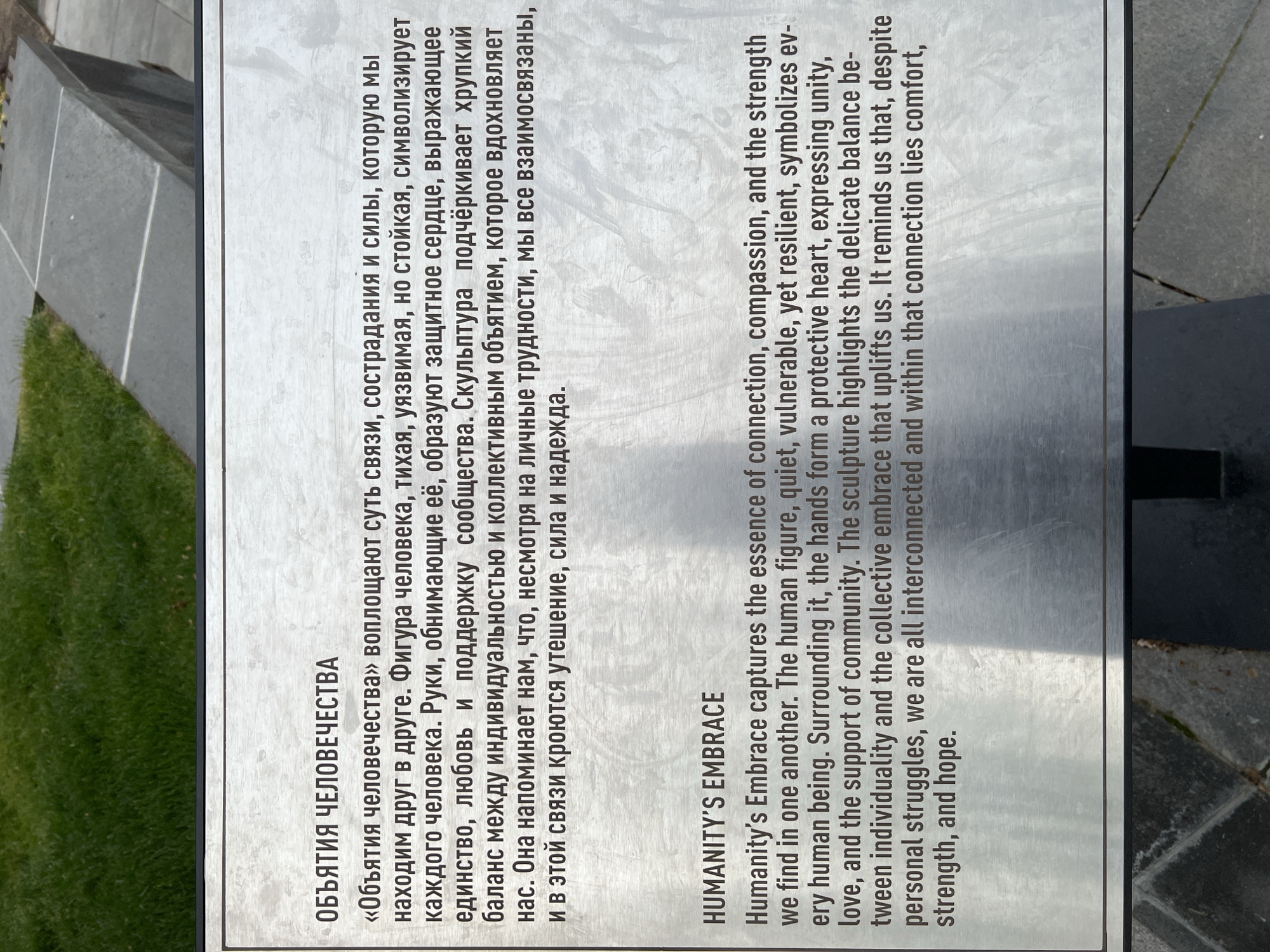 The image shows a metal sign with text in Russian and English. The text describes the sculpture "Humanity's Embrace", which symbolizes the essence of connection, compassion, and strength found in one another. The Russian text is a translation of the English. The sign is located in Krasnodar, Russia, near the "83rd Border Guards" memorial in the city park.