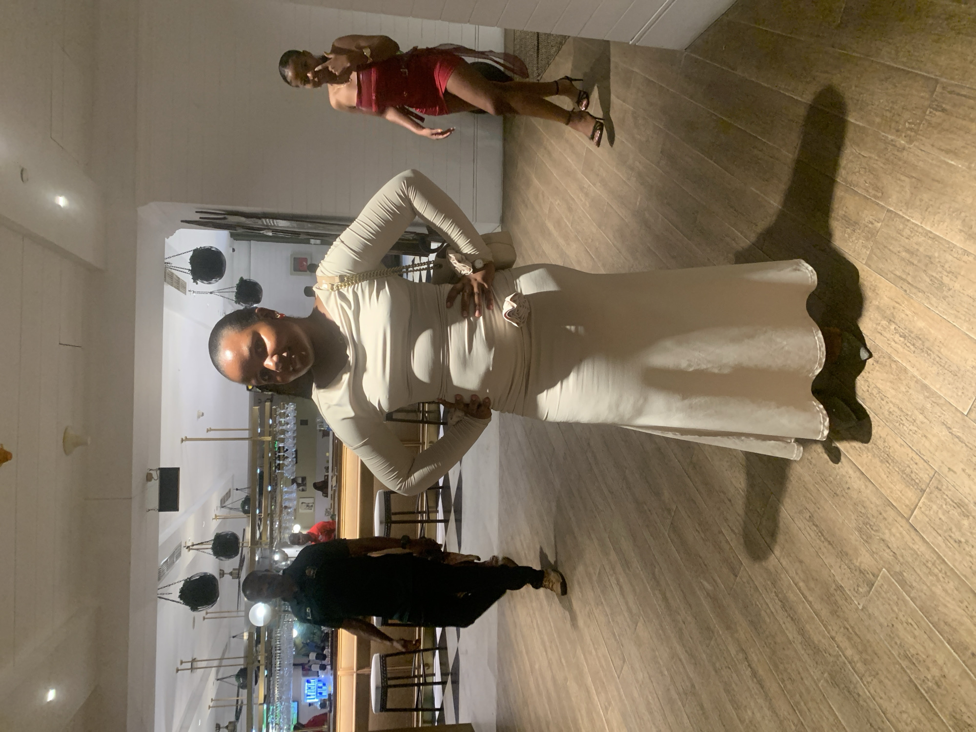 The image shows two women indoors, possibly in a bar or restaurant setting. One woman is in the foreground, posing in a long cream-colored dress. The other woman is in the background, wearing a short red dress and heels. In the background, there is a bar with chairs and what appears to be a bartender standing behind it. The interior features wooden floors, hanging light fixtures, and white walls. The location is likely in Dar es-Salaam, Tanzania, due to the person's prompt request.