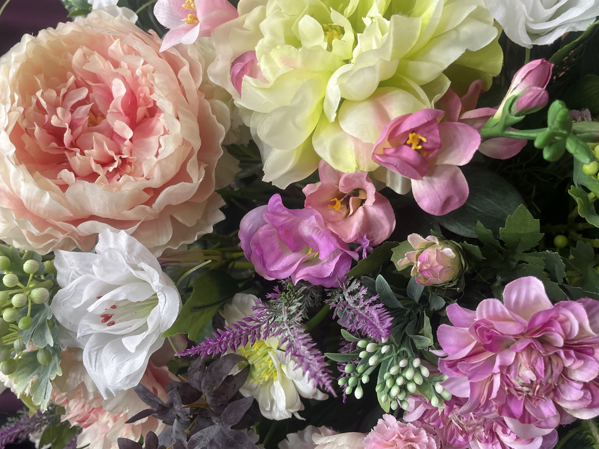 This close-up image showcases a vibrant and dense arrangement of artificial flowers, filling the entire frame. The subjects are various types of faux blossoms and foliage, presenting a harmonious blend of pastel and jewel tones.
A prominent feature is a large, fully-bloomed light pink and peach peony-like flower positioned on the left side, characterized by its numerous ruffled petals. Towards the top center, a large, multi-layered flower with light yellow-green petals, possibly resembling a dahlia or chrysanthemum, stands out. Below the peony, a white lily-like flower with delicate petals and visible red markings or stamens inside adds contrast.
Smaller pink and purple flowers are interspersed throughout the arrangement, including simple pink blossoms with bright yellow centers, bud clusters, and larger, multi-petaled purple flowers, similar to asters or dahlias, visible on the lower right. Complementing the blooms are various green leaves, dark purple foliage, and spike-like purple accents that mimic lavender or heather. Clusters of small, round, light green berry-like elements are also visible, adding texture.
No activity or event is taking place; the image is a static portrayal of the floral display. The lighting is bright and even, accentuating the colors and fabric textures of the artificial flowers. The background is indistinct and dark, ensuring the focus remains entirely on the detailed arrangement. There is no visible text in the image, and no specific time of day or weather can be discerned. The composition suggests an abundant and decorative floral piece, evoking a sense of beauty and tranquility. No location cues are visible within the image itself.