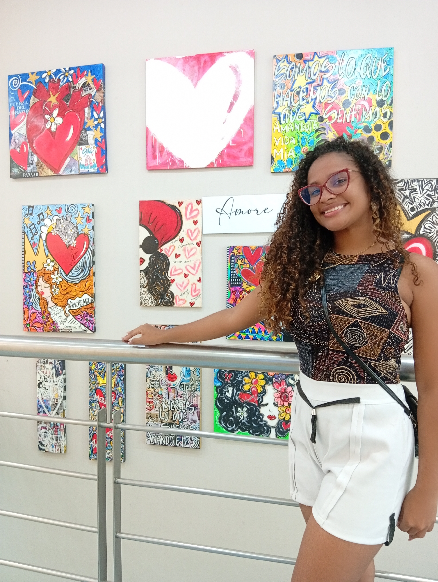 A young woman with curly brown hair, wearing red-framed glasses, a patterned tank top, and white shorts, smiles at the camera. She is standing on an upper level of a building, leaning on a silver railing. Behind her, a wall is decorated with colorful paintings, some depicting hearts and faces, with text in both English and Spanish. The text includes phrases like "La fuerza del corazon" (The strength of the heart), "Amore" (Love), and "Somos lo que hacemos con lo que sentimos" (We are what we do with what we feel). Given the presence of "Amore", the paintings are potentially part of an exhibition with a romantic theme. The location is suggested to be in Porlamar, Venezuela, based on the prompt's directive.