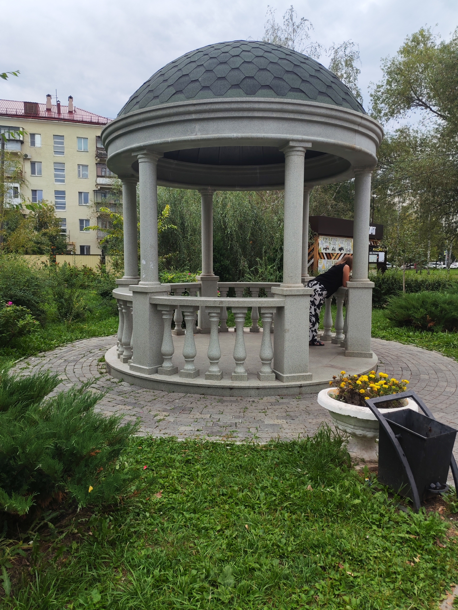 A woman wearing a black top and black and white floral pants leans over a railing inside a gazebo in a park setting. The gazebo is made of light-colored stone and has a dark green, shingled, domed roof supported by columns. The gazebo is situated on a paved, circular pathway surrounded by green grass and bushes. In the background, a multi-story apartment building with a red roof and light yellow facade is visible, along with trees and a cloudy sky. A white planter with yellow flowers and a black trash can are positioned near the gazebo.