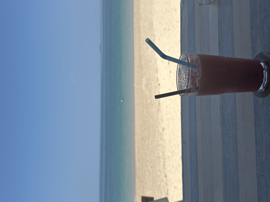 A tall, transparent glass containing a red-pink liquid with external condensation rests on a dark coaster atop a multi-planked wooden surface in the foreground. Two straws, one dark and straight, one light blue and bent, protrude from the liquid. The wooden surface consists of alternating light blue and light gray horizontal planks. A small, clear glass ashtray is partially visible to the right. The midground presents an expansive, unoccupied light-tan sandy beach. The background features the Andaman Sea, exhibiting a gradient from light turquoise near the shore to a darker blue further out. A single white buoy floats in the water towards the middle-left. A small, dark landmass is discernible on the distant horizon, towards the right. The upper sky is clear and uniformly blue. This scene is situated in Phra Ae, Thailand.
