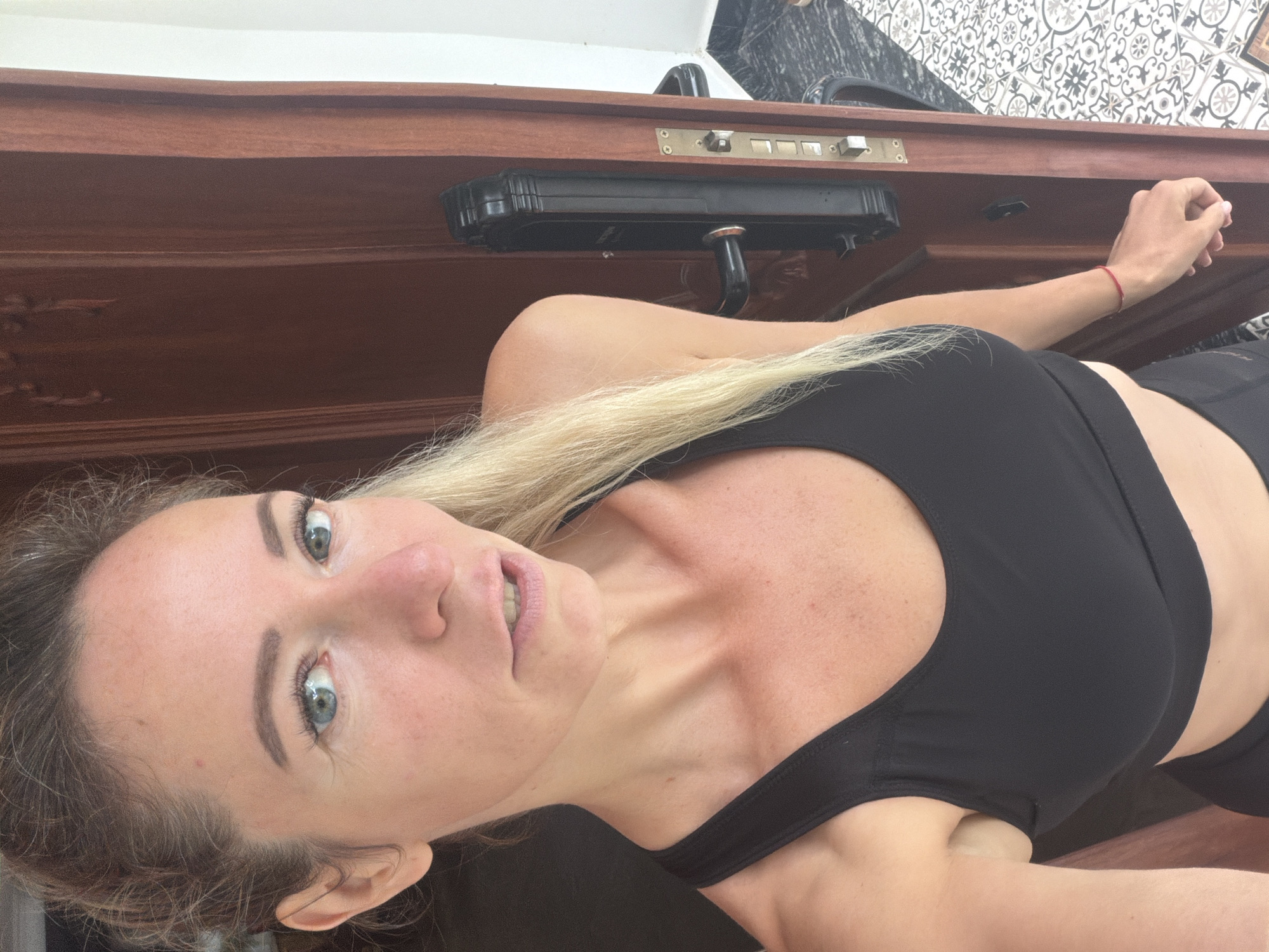 A woman stands in a doorway, her body angled towards the viewer. She has blonde hair pulled back in a ponytail, and her face is turned slightly to the side, with her mouth open in a slight smile. She is wearing a black sports bra and dark bottoms. The doorway is made of dark wood, and a black door handle is visible on the right side. The floor is tiled with a geometric pattern. The scene appears to be outdoors, as indicated by the natural light. The woman's pose and attire suggest she may be engaged in or has just finished physical activity. The location is identified as Xuân Trường - Đà Lạt, Vietnam.