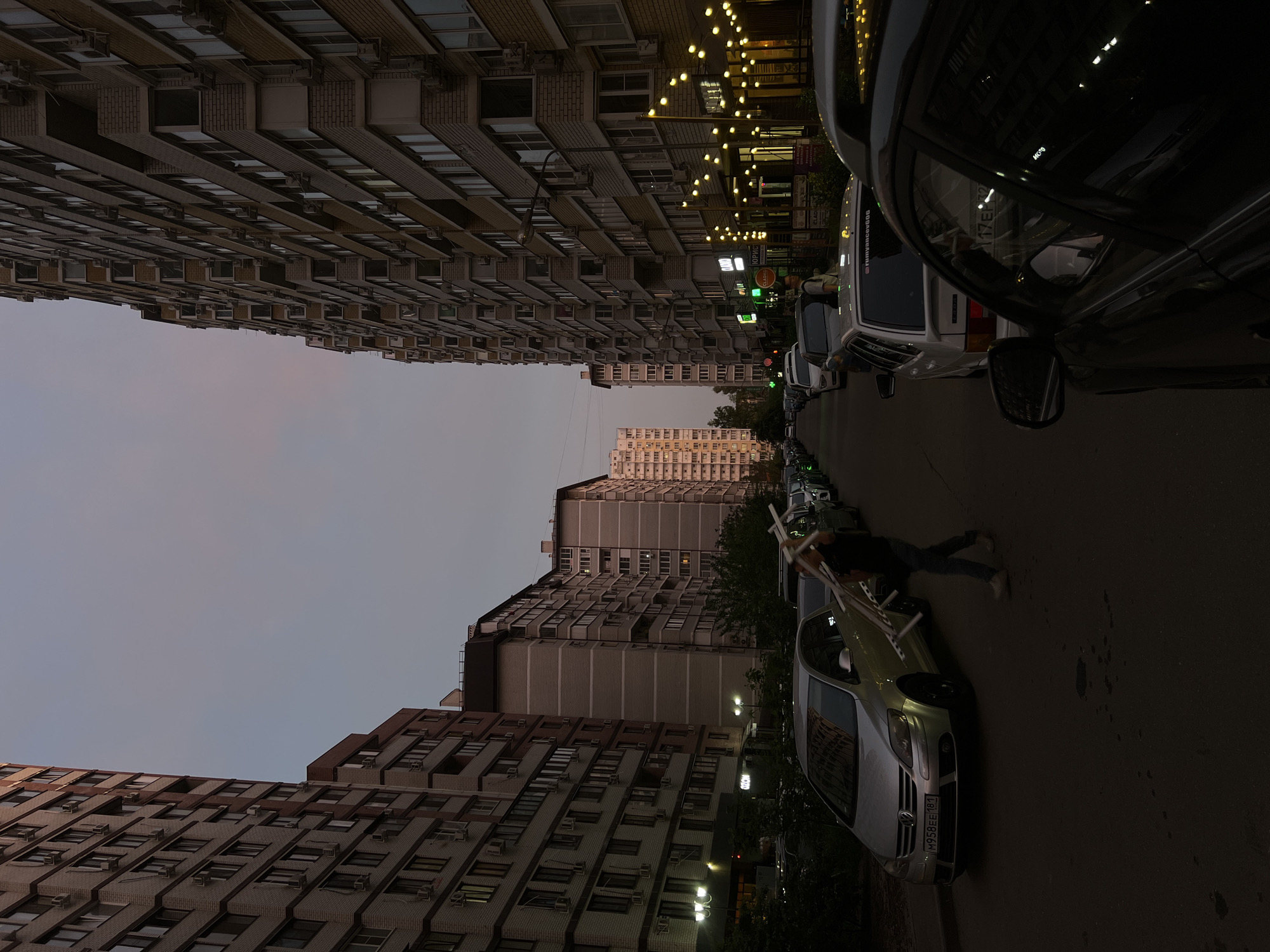 The image shows a street in Krasnodar, Russia, lined with tall apartment buildings on both sides. Cars are parked along the street. A person is carrying a white object across the street between the parked cars. The sky is dusky, suggesting twilight. Streetlights are visible and illuminated. The buildings are densely packed with many windows and air conditioning units.