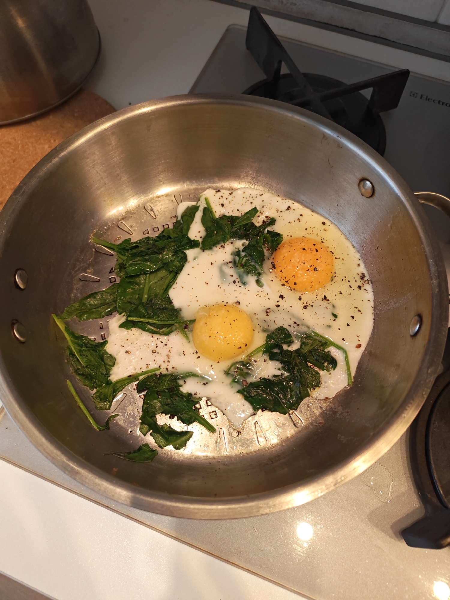 In a silver frying pan on a stovetop, two fried eggs with visible yolks are nestled amongst wilted spinach leaves. The eggs and spinach are seasoned with black pepper. The pan is slightly greasy and sits on a white countertop next to a gas burner. A metallic pot is visible in the upper left corner of the image.