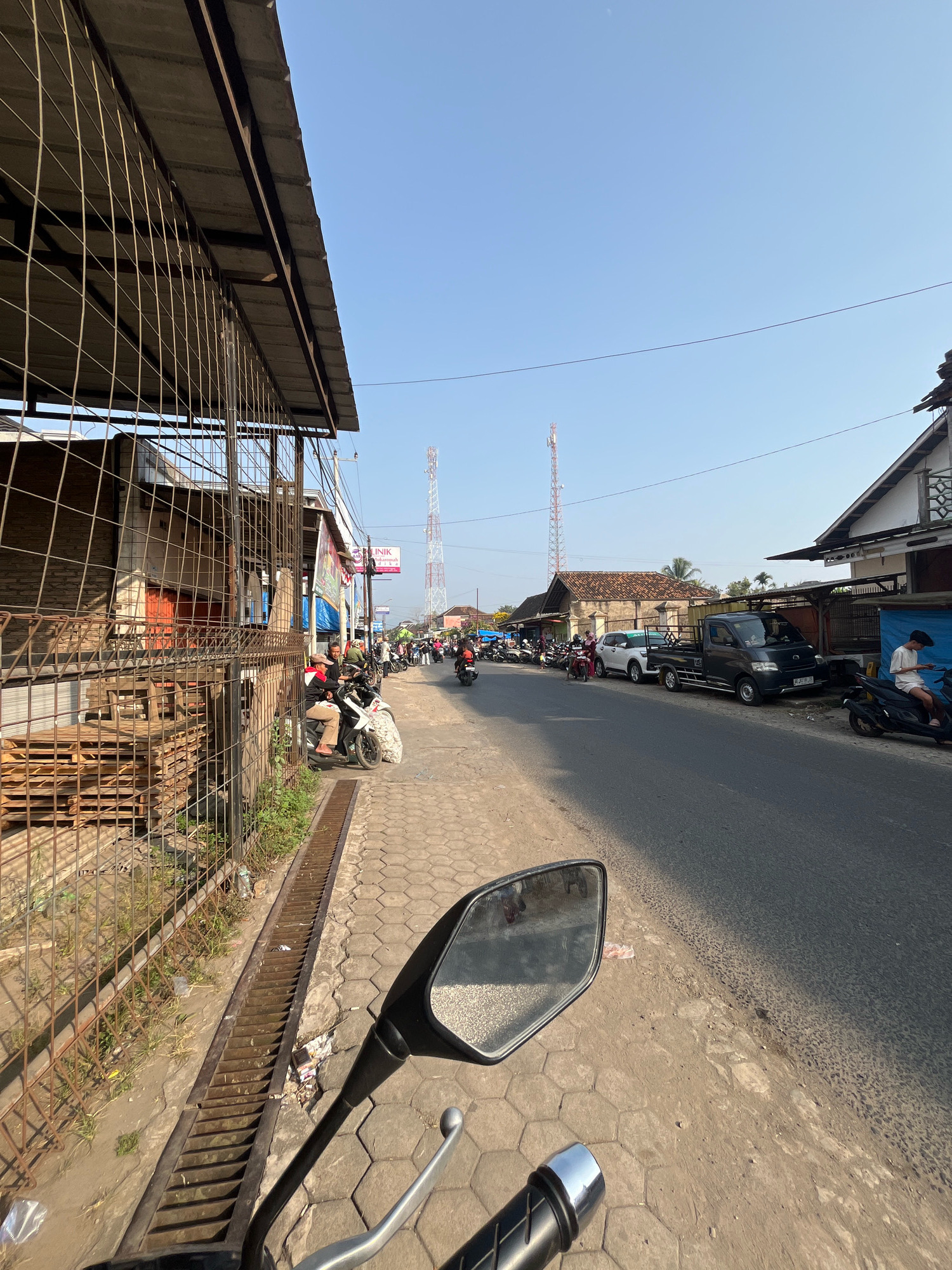 The image depicts a street scene in Kemuning, Indonesia. On the left, a weathered metal fence encloses an area with stacked wooden pallets. A paved sidewalk runs alongside the street, with a metal grate drain running parallel to the fence. Several people on motorcycles are parked or passing by on the sidewalk and street. A dark gray pickup truck and a white SUV are parked on the right side of the street. In the background, buildings with tiled roofs line the street, and two prominent cellular towers are visible against a clear blue sky. The image appears to have been taken from the perspective of someone on a motorcycle, with the motorcycle's mirror and handlebars visible in the foreground.