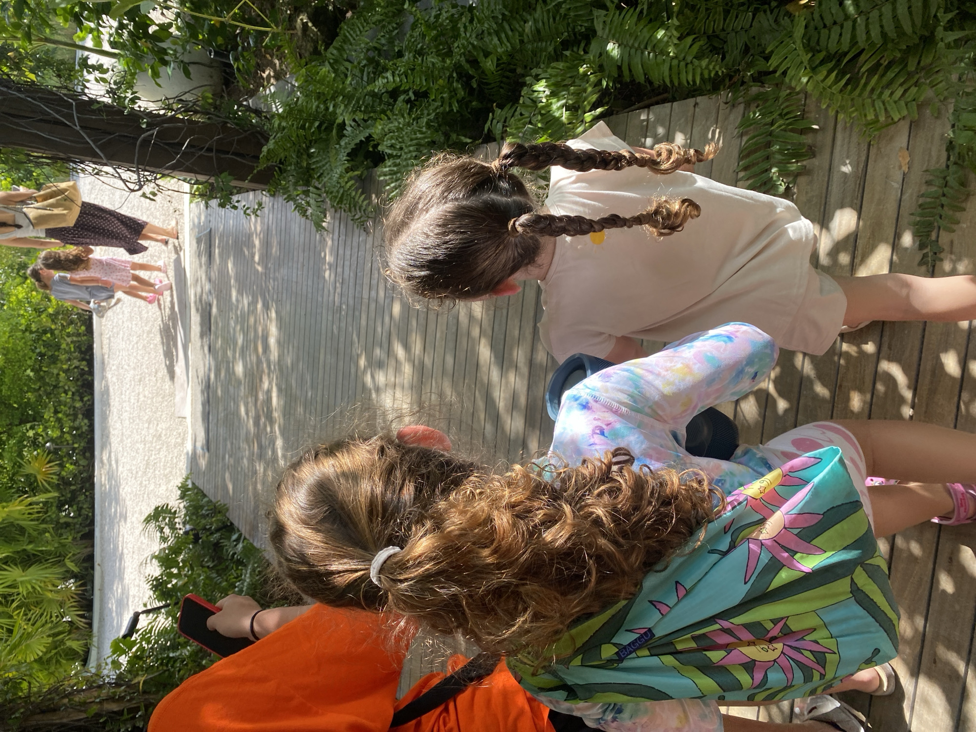 The image shows an outdoor scene with a wooden walkway. Two young girls are in the foreground, one with braids and wearing a light-colored shirt, and the other with curly hair tied in a ponytail, wearing a colorful shirt and carrying a reusable shopping bag with a floral pattern. An adult wearing an orange shirt is holding a phone. In the background, other people can be seen walking on the path. Lush greenery surrounds the walkway. The location is likely Marina Maroma, Mexico, based on the user's prompt.
