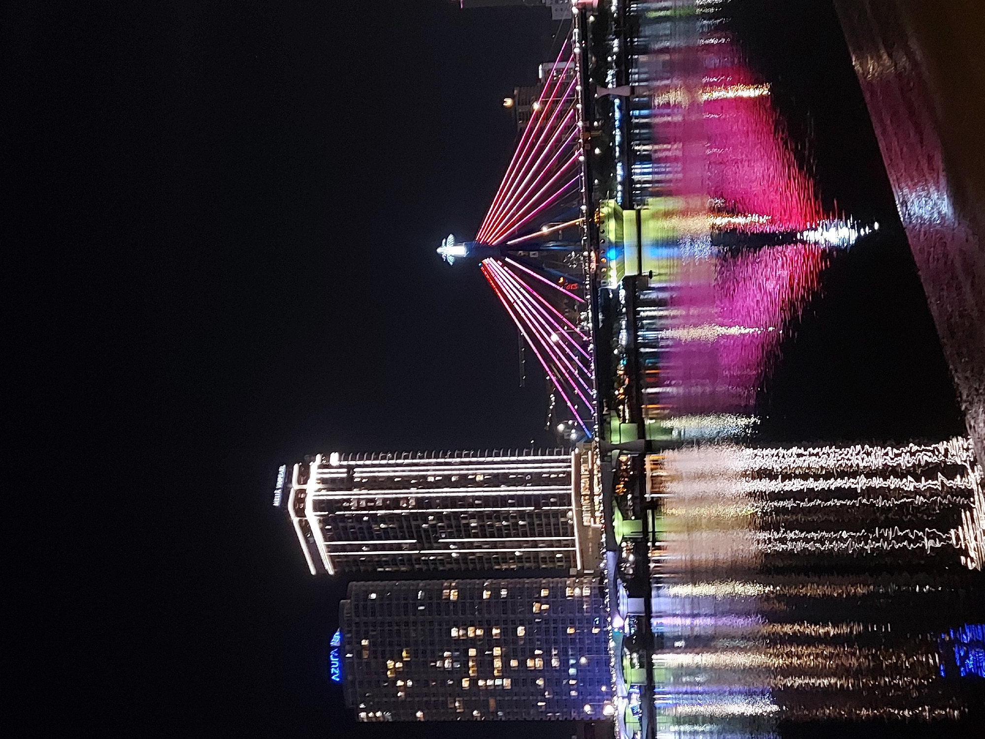 The image depicts a nighttime view of the Dragon Bridge in Đà Nẵng, Vietnam. The bridge is illuminated with vibrant pink and red lights that stretch across its span, creating a dramatic visual effect against the dark sky. A tall tower at the center of the bridge is topped with a cross-shaped structure, also illuminated. In the background, two prominent skyscrapers are visible. The skyscraper on the left is labeled "AZURA" in blue neon lights, and the one on the right is labeled "MELIA VINPEARL" in white neon lights. Both buildings are lit up with numerous windows. The bridge and buildings are reflected in the dark water of the Han River, with streaks of light shimmering on the surface. The foreground shows a dark, wet surface, possibly a walkway or quay. The overall scene is a cityscape at night, characterized by bright artificial lights and their reflections.