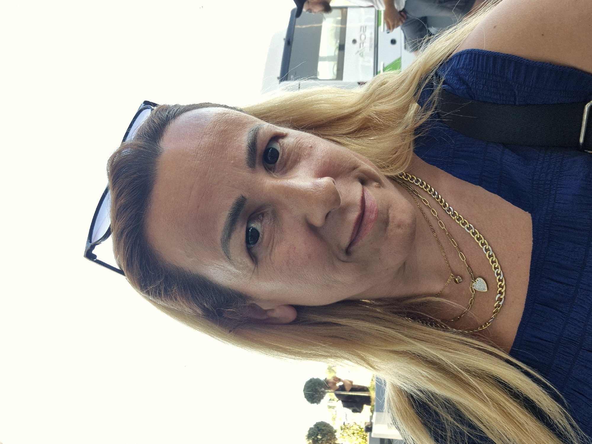 This is a close-up, vertically oriented, portrait-style photograph of a woman. The woman has blonde hair, brown eyes, and fair skin with some freckles. She is wearing a blue, textured dress and several gold necklaces, one of which has a heart-shaped pendant with small stones. She is also wearing dark-rimmed glasses, which are perched on her head.
The background is out of focus and suggests an outdoor setting. A white vehicle with green and black accents is visible, with text that appears to read "ARDE". Two blurred figures can be seen in the distance, one in a dark shirt and another in a lighter shirt. The lighting suggests it is daytime, possibly sunny, given the brightness of the background. The woman's expression is a slight smile, conveying a relaxed and pleasant emotion. The orientation of the photo suggests it was taken by the woman herself, likely a selfie.