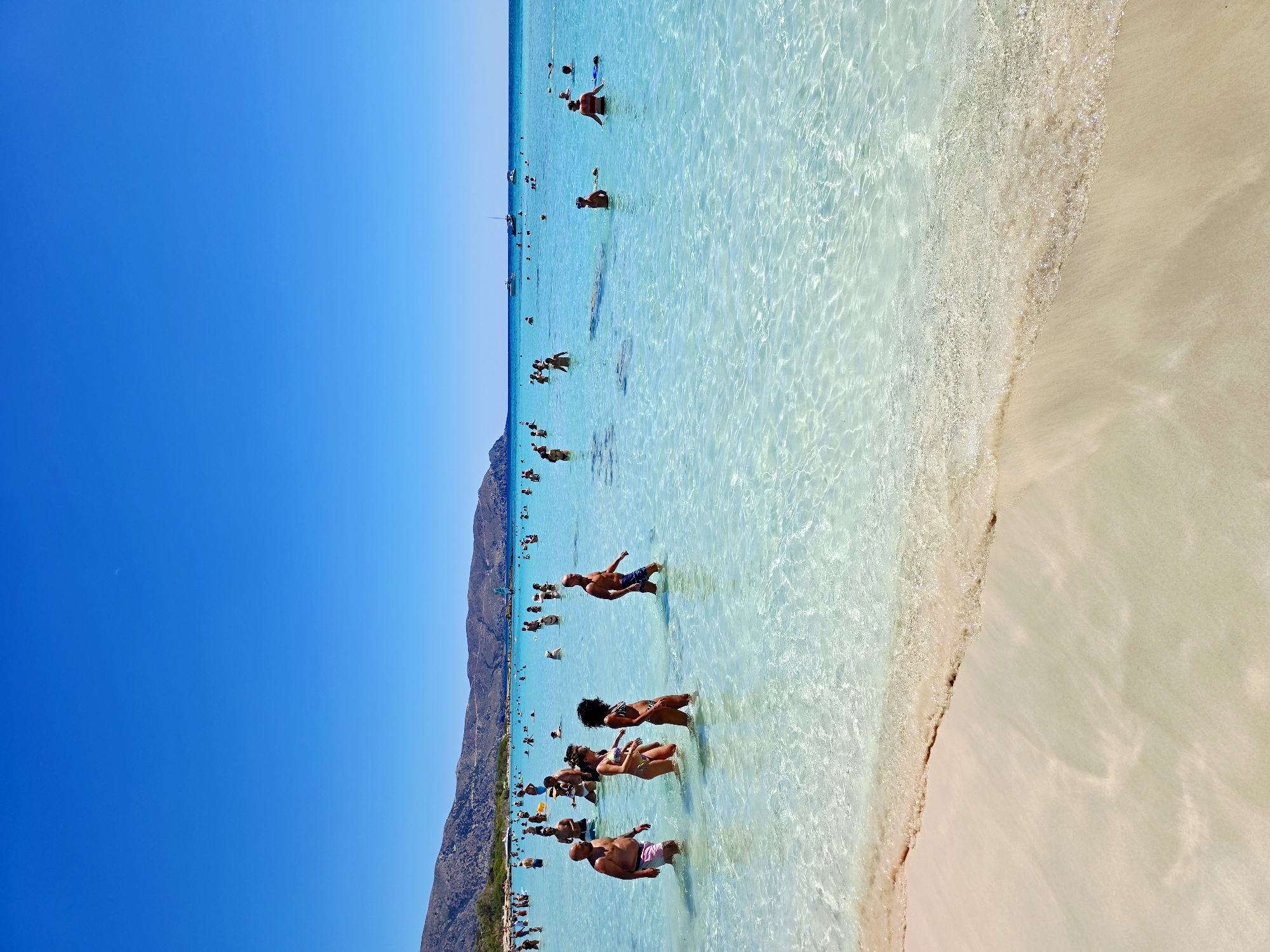 The image depicts a crowded beach scene with numerous people wading in the shallow, crystal-clear turquoise water under a bright blue sky. In the foreground, a gentle wave laps onto a light-colored sandy shore. The water is so transparent that the sandy bottom is visible. A group of individuals are scattered throughout the water, some appearing to be swimming or standing, while others are walking through the shallows.
Further out, more people are visible, and a few boats are anchored or sailing on the horizon. In the background, a chain of low, arid hills with sparse vegetation forms a backdrop to the beach. The overall impression is one of a popular summer destination with abundant sunshine and activity. The location is identified as Elafonissi Beach, Crete, Greece.