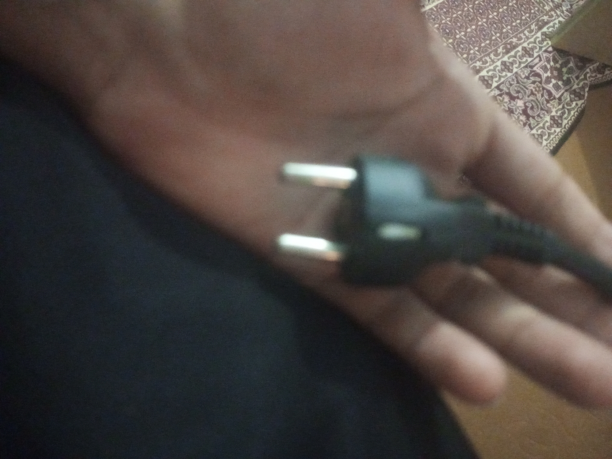 A close-up, blurry image shows a hand holding a black electrical plug with two prongs. The background is dark, with a patterned rug visible on the right. The location in Mansehra, Pakistan is not discernible from the image.