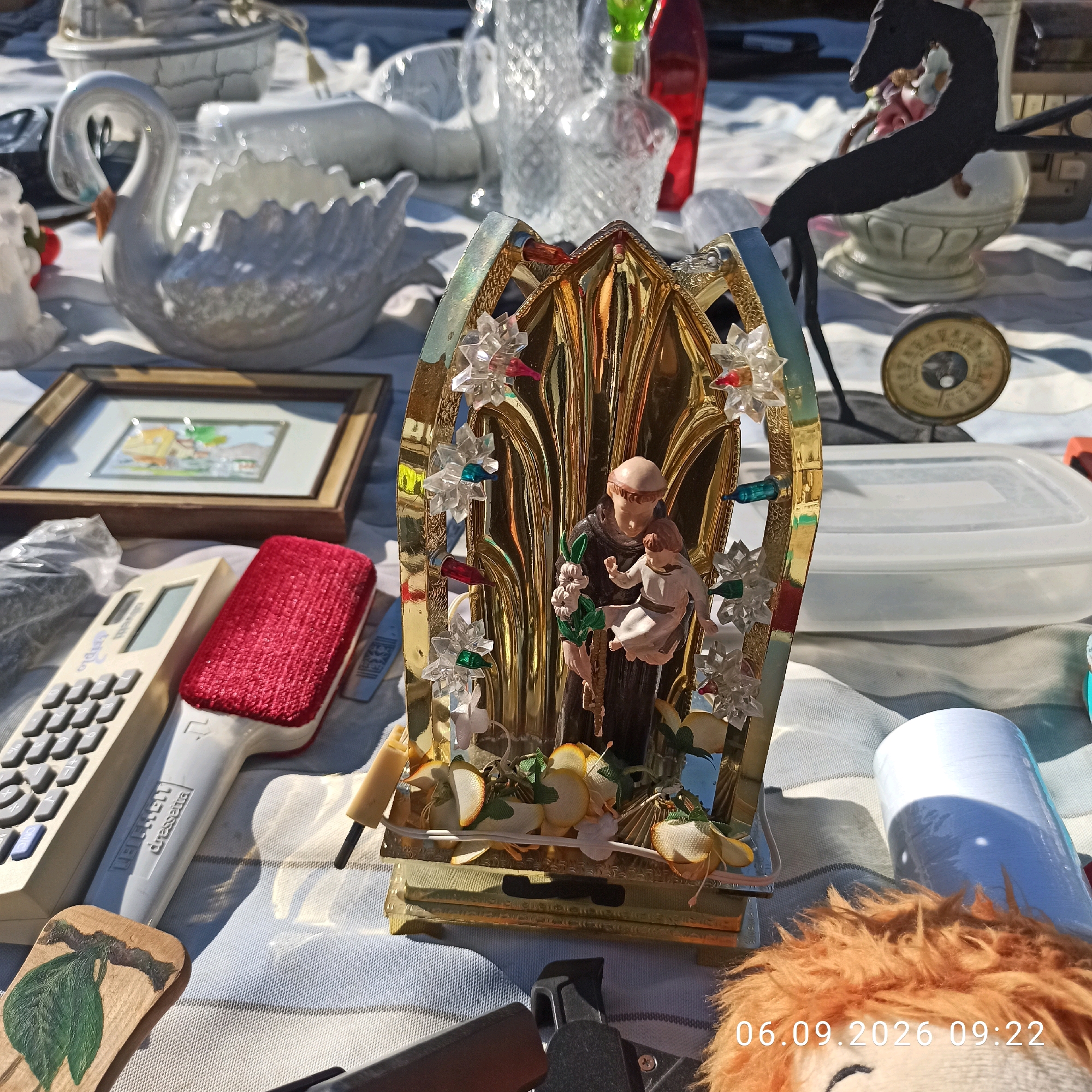 The image depicts an outdoor market stall, likely a flea market, with various items laid out on a striped cloth. In the foreground, a beige calculator and a red-bristled cleaning tool are visible. To the left, a small wooden plaque painted with a green leaf hangs by a string. A decorative religious figurine featuring Saint Anthony holding a child is prominently displayed in the center, encased in a gold-colored ornate frame adorned with clear and colored acrylic "jewels" and artificial flowers. Behind the figurine, a framed picture of a house is visible. Further back, a white ceramic swan is partially visible, along with other household items and decorative pieces including a glass decanter and a dark metal sculpture of a horse-like figure. A small circular gauge or clock with a dial is also present. In the lower right corner, a plush toy with orange shaggy hair is partially in view, and a timestamp "06.09.2026 09:22" is superimposed on the image. The lighting suggests it is daytime, with shadows indicating sunlight. The presence of various vintage and decorative items suggests a setting for secondhand goods. The timestamp and overall presentation are consistent with a street market or similar outdoor sales event. The city of Sannicandro di Bari, Italy, is known for its historical sites and agricultural landscape, but the image itself does not contain specific visual cues to definitively place it within this exact location.
