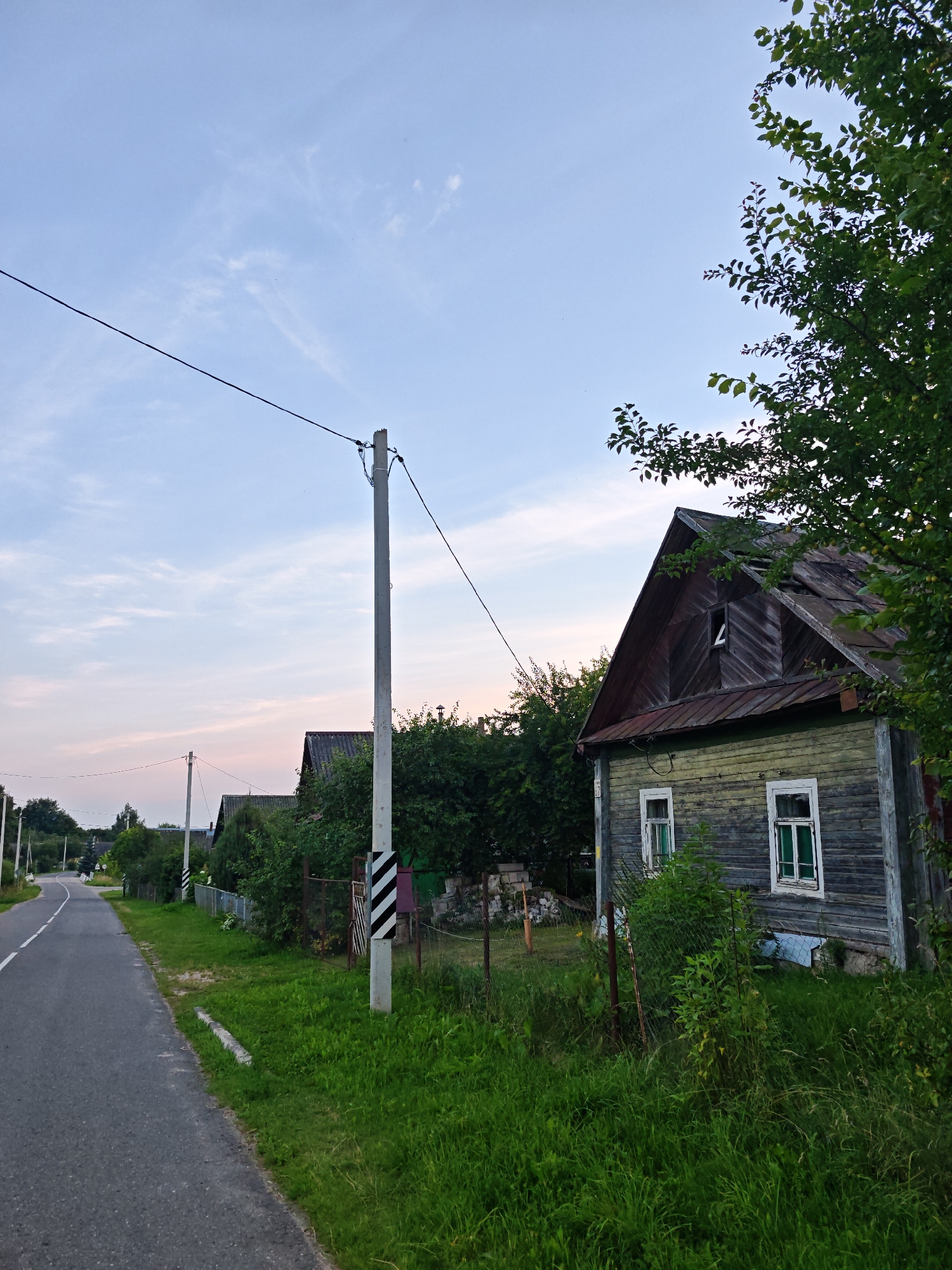 The image depicts a rural scene in the Валожынскі сельскі Савет, Belarus. A paved road curves to the left, bordered by a grassy verge. On the right side of the road, an old, weathered wooden house stands with a dilapidated roof. The house has two visible windows with white frames and green curtains. Adjacent to the house is a wire fence overgrown with vegetation. A concrete utility pole with black and white diagonal markings stands near the road, supporting electrical wires that stretch across the frame. In the background, more houses and lush green trees are visible under a pale, cloudy sky. The overall atmosphere is quiet and suggests a late afternoon or early evening setting. No people are present in the image.