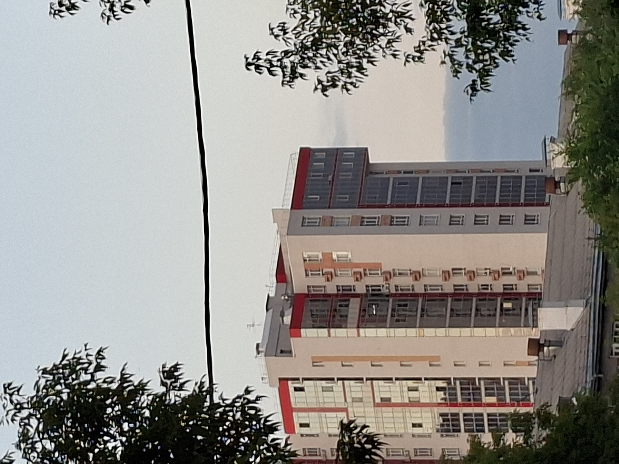 The image shows a residential building in Kazan, Russia. The building is tall and modern, with a light-colored facade and red accents on the upper floors. The windows are a mix of clear glass and reflective panels. There are trees in the foreground and background, obscuring parts of the building. The sky is overcast.